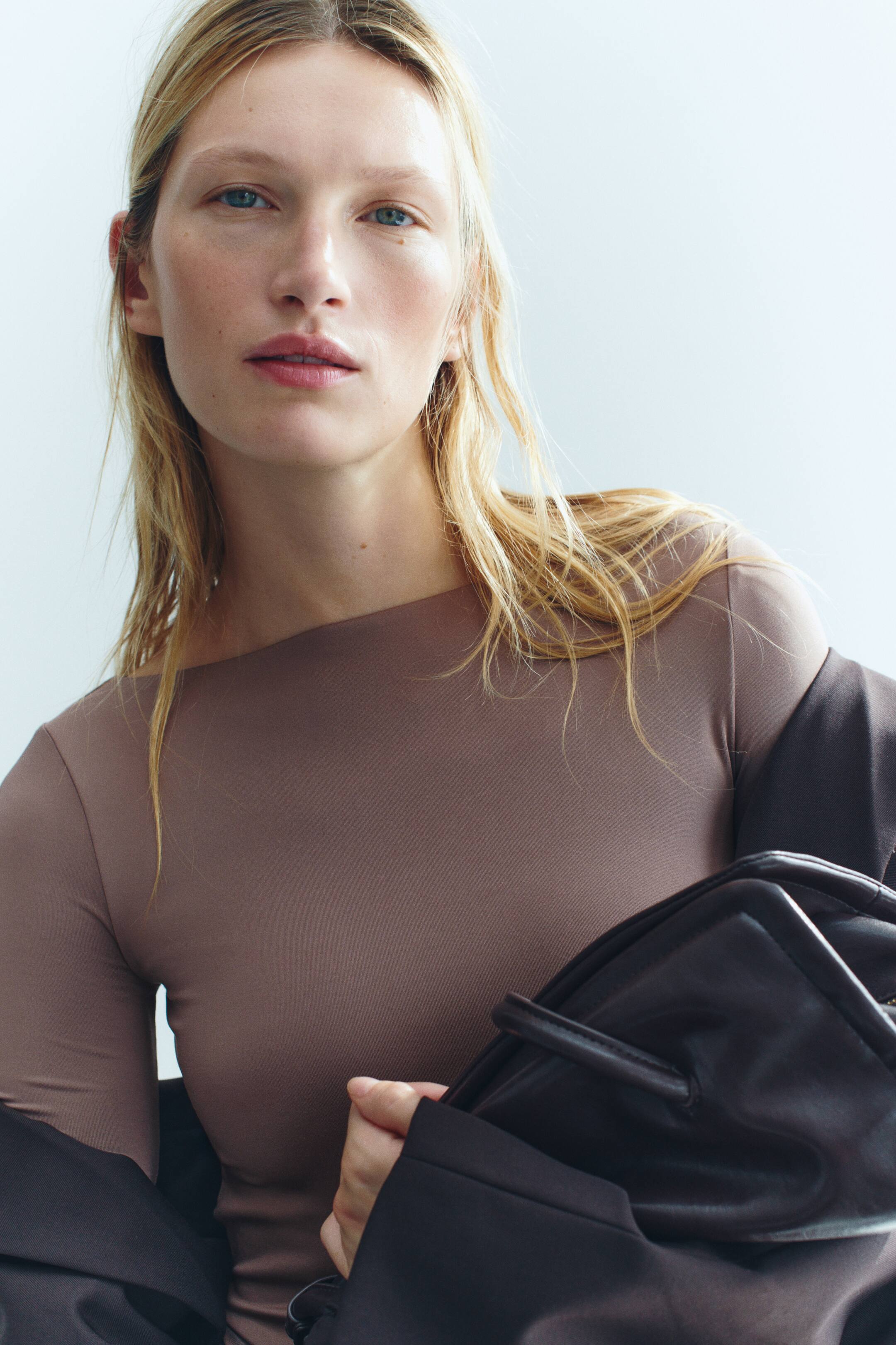 View larger image: The model is wearing a fitted taupe stretch jersey top with a boat neck. A dark jacket is draped off the shoulders over the top, and a dark leather bag is being held in front.