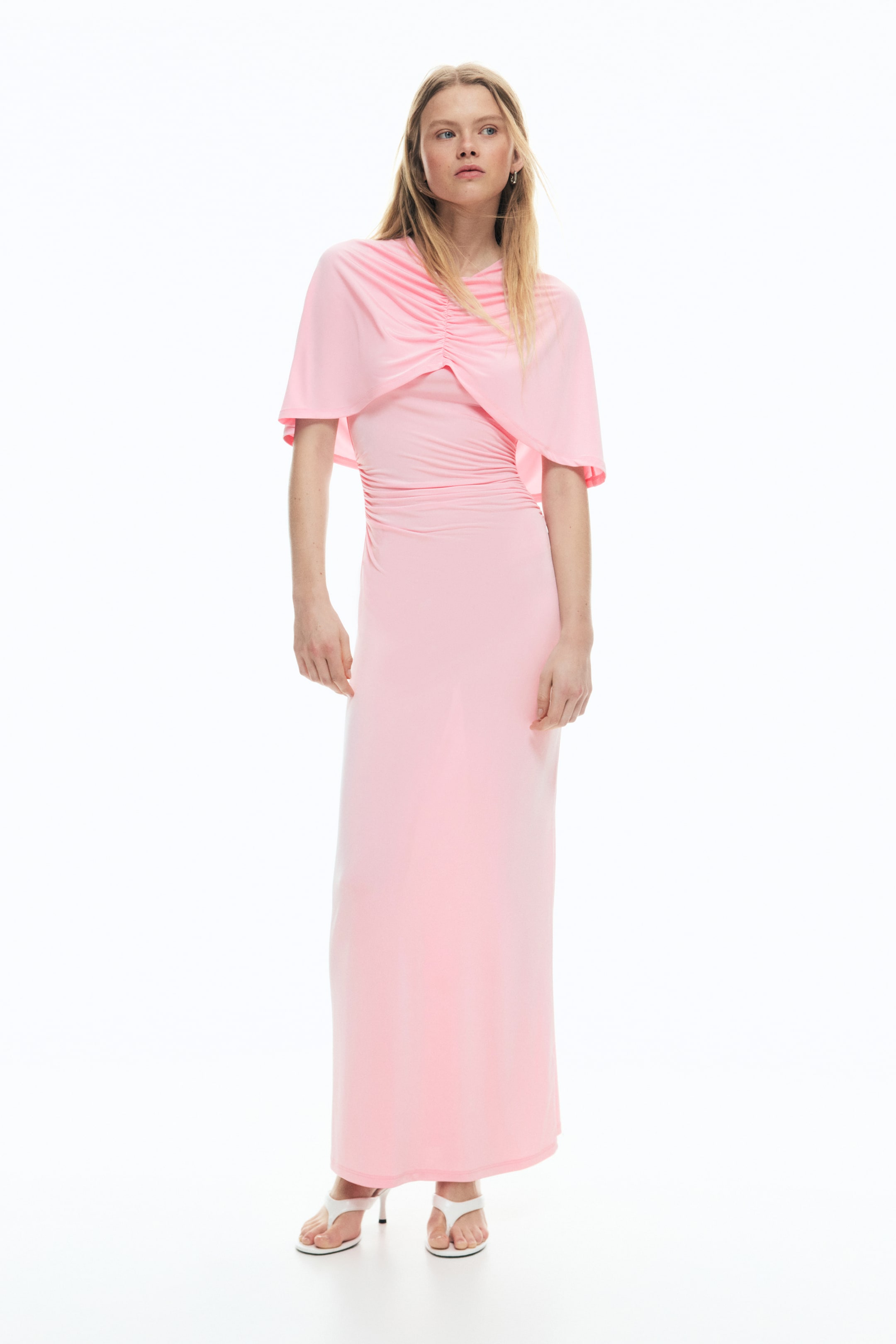 View larger image: The model is wearing a pink calf-length jersey dress. It features a round neckline, a cape-like layer over the top with a gathered seam at the front, and gathers that create a draped effect at the waist.