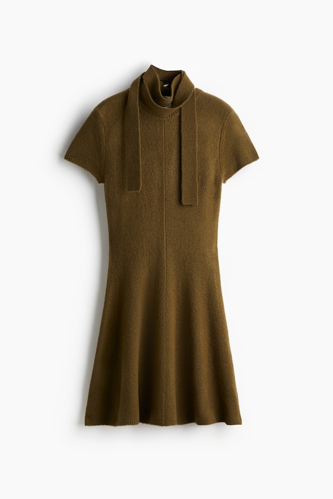 Une robe en tricot texturé d'un vert olive chaud est présentée de face, avec un col montant haut et deux longues attaches en tissu plat tombant librement sur le devant. Elle est conçue avec des manches courtes et une silhouette en ligne A qui s'évase doucement jusqu'à un ourlet au-dessus du genou. - 1