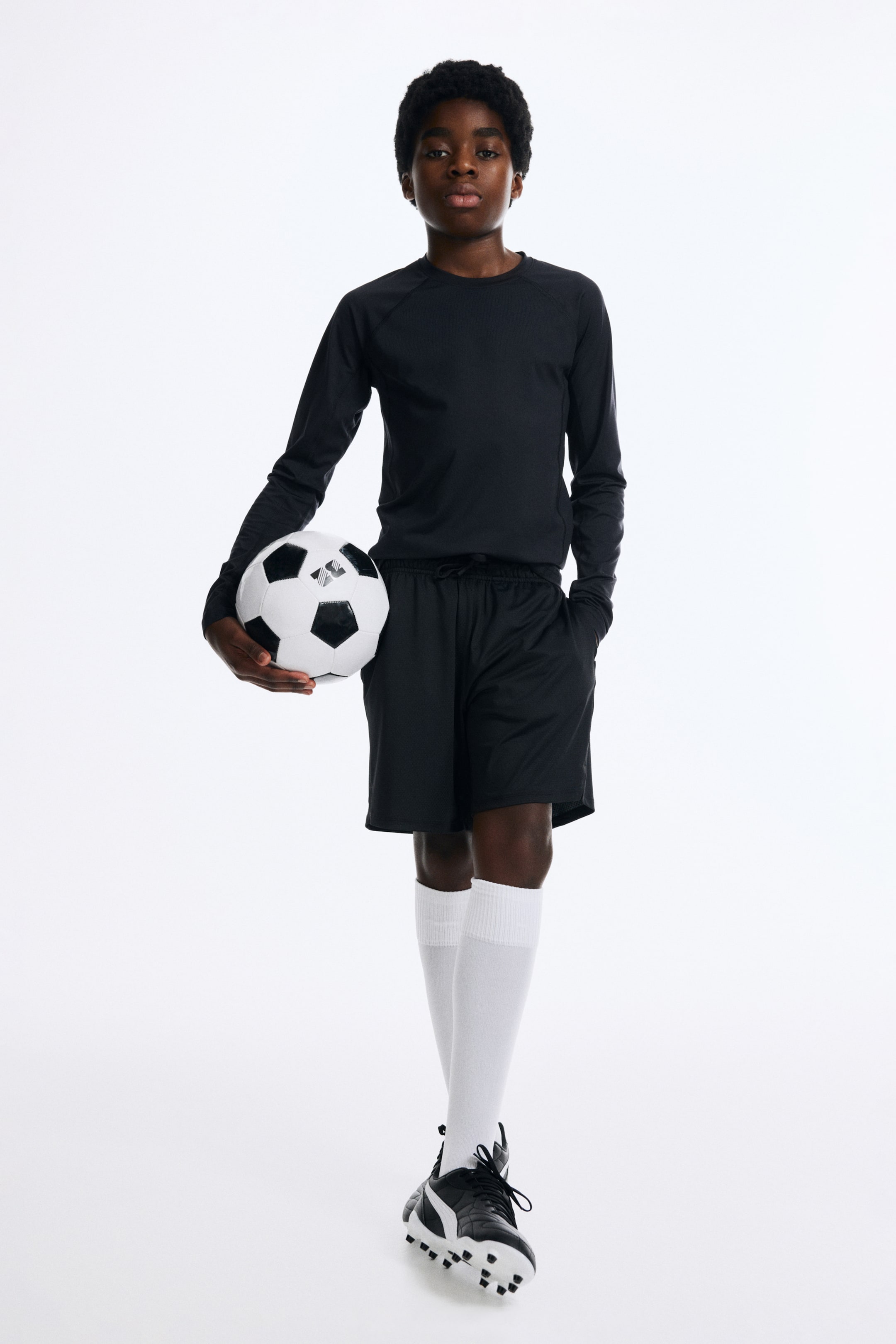 Näytä suurempi kuva: Musta pitkähihainen urheilupaita raglanhihoilla ja hienovaraisesti teksturoidulla kankaalla, mustat urheilushortsit kiristysnauhallisella vyötäröllä ja verkkomaisella pinnalla, valkoiset polvenkorkuiset urheilusukat resorireunuksin sekä mustavalkoiset jalkapallokengät selkeästi erottuvilla nastoilla esitetään edestä päin. Mustavalkoinen jalkapallo pidetään kädessä.