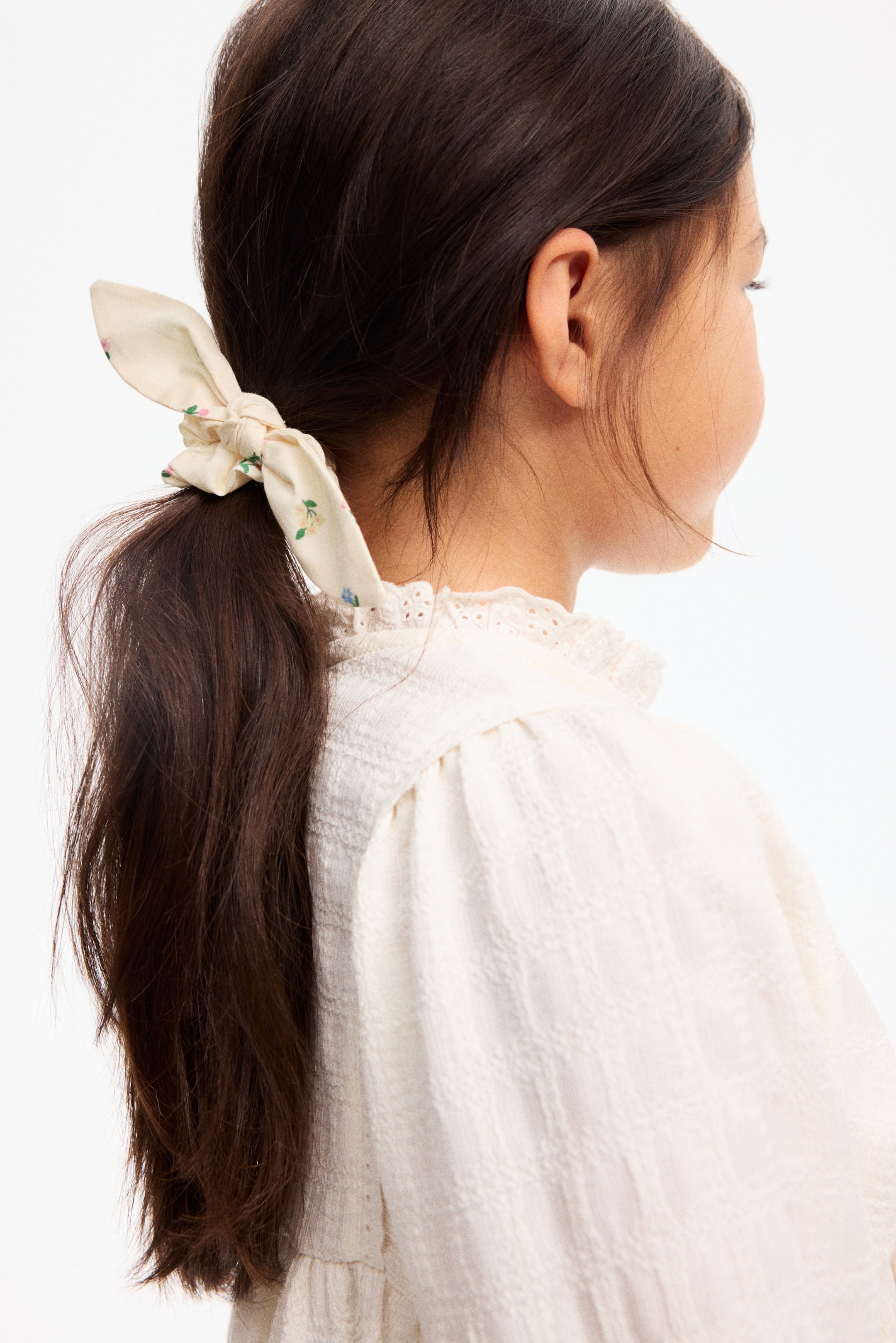 View larger image: A cream-colored scrunchie with a small floral print secures a ponytail. The scrunchie features a tied bow detail with two short fabric tails extending upward.