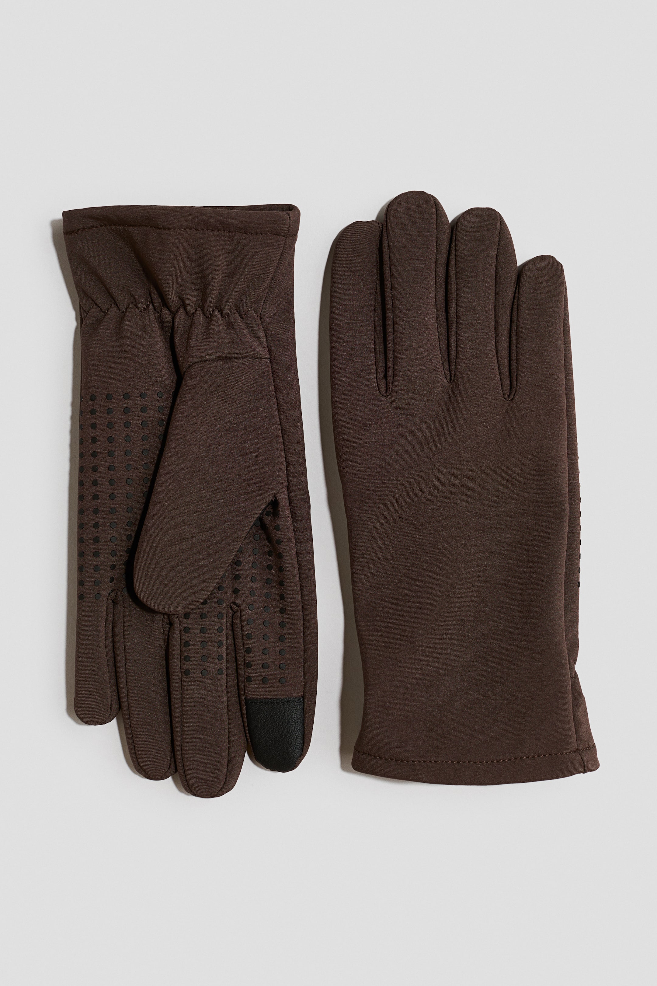 View larger image: A pair of dark brown gloves is laid flat, showing both the palm side and the back of the hand. The palm side features an elasticated wrist, black anti-slip grip dots, and a black touchscreen-compatible index fingertip, while the back has a smooth fabric finish.