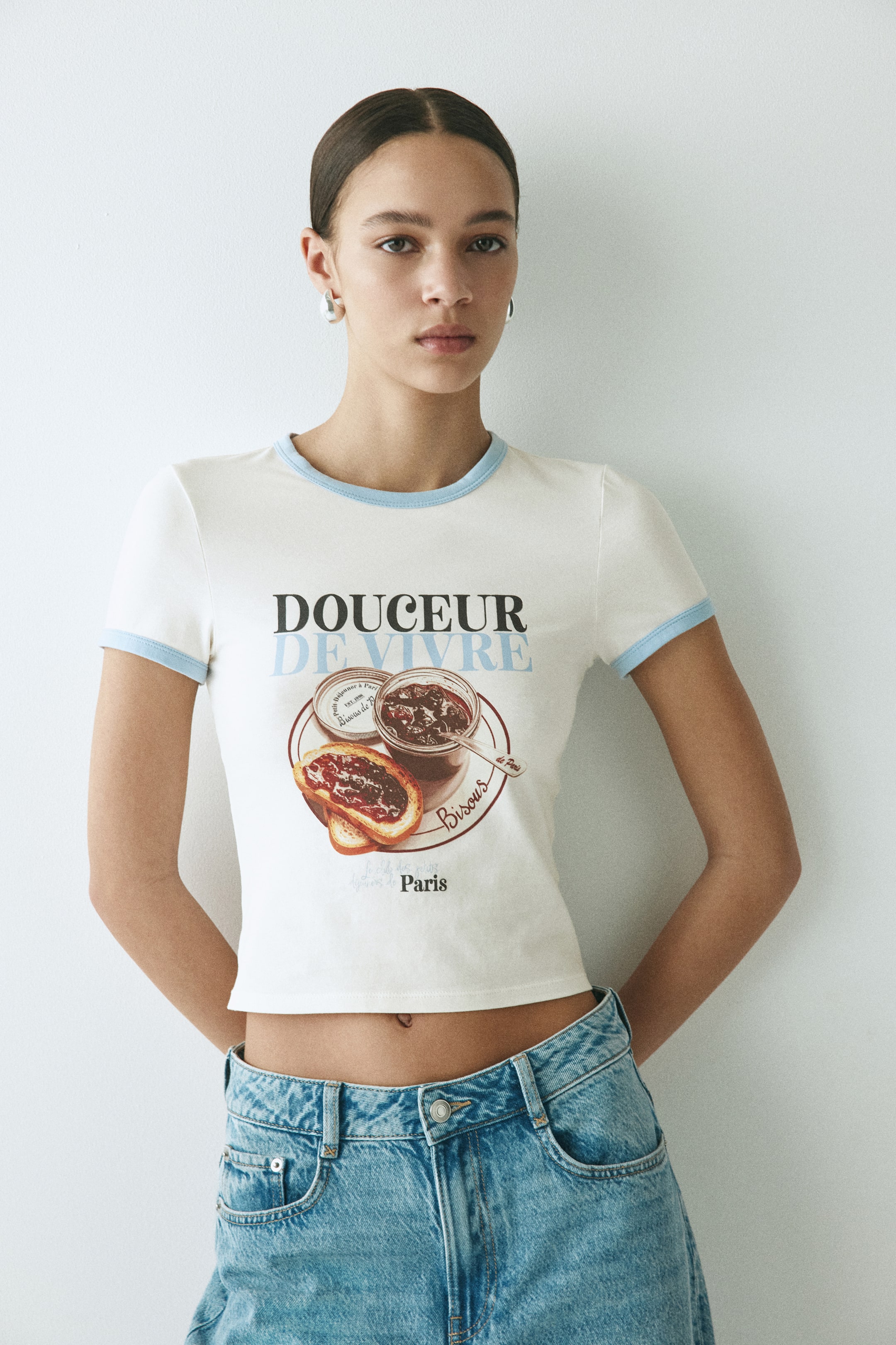 Ampliar la imagen: Playera ringer en blanco roto que presenta una silueta cropped y ceñida, con ribetes azul claro en el cuello tipo "crew" (redondo) y en los puños de las mangas cortas. Un gráfico central muestra el texto en negro y azul claro "DOUCEUR DE VIVRE" sobre una ilustración de un frasco de mermelada y ésta sobre una rebanada de pan, rodeado por los textos "Bisous" y "Paris".