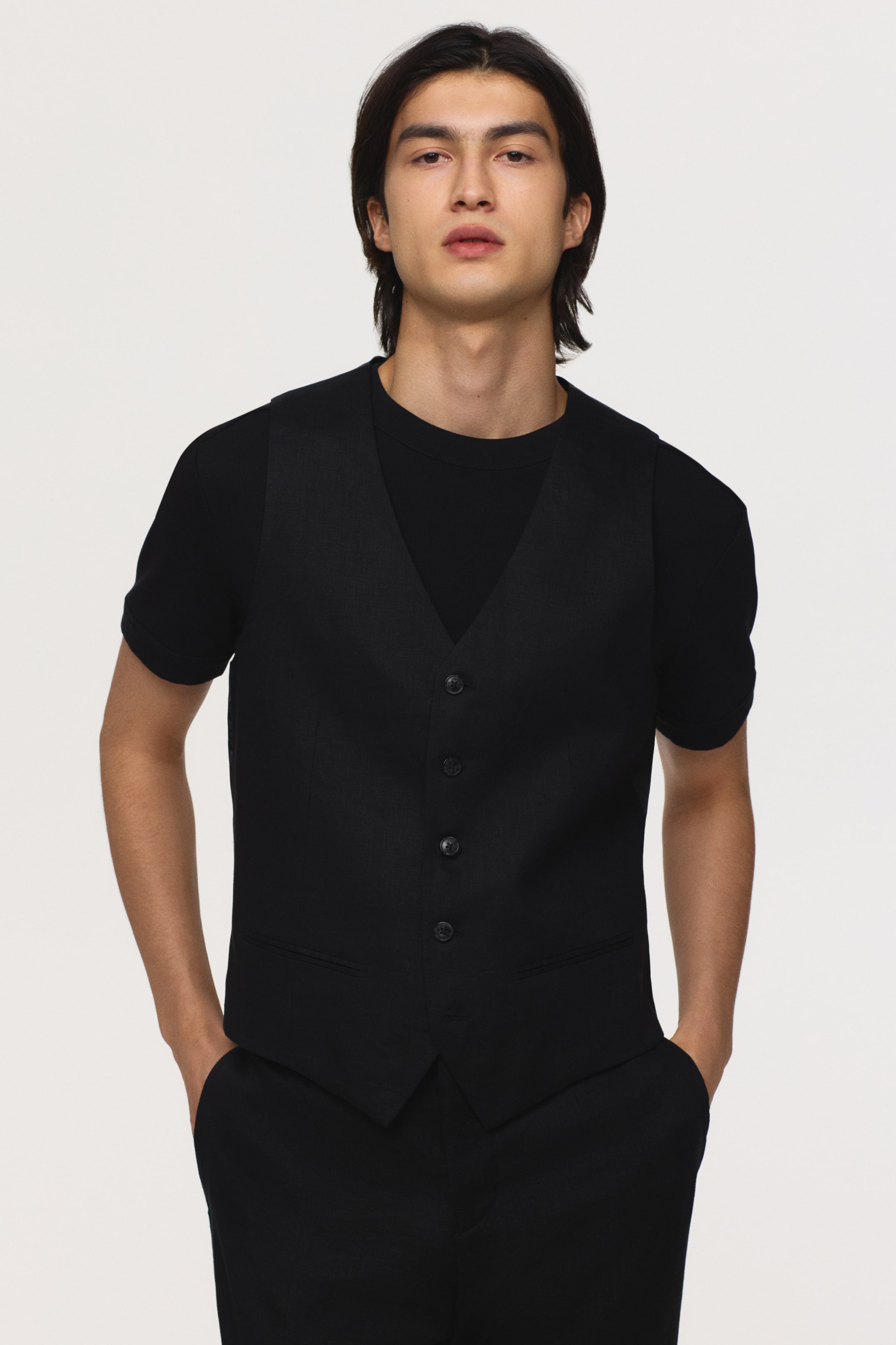 View larger image: Introducing a sleek black waistcoat, showcasing a V-neckline and five visible buttons running down the front. It features two small welt pockets near the hem, perfectly styled over a black crew-neck t-shirt.