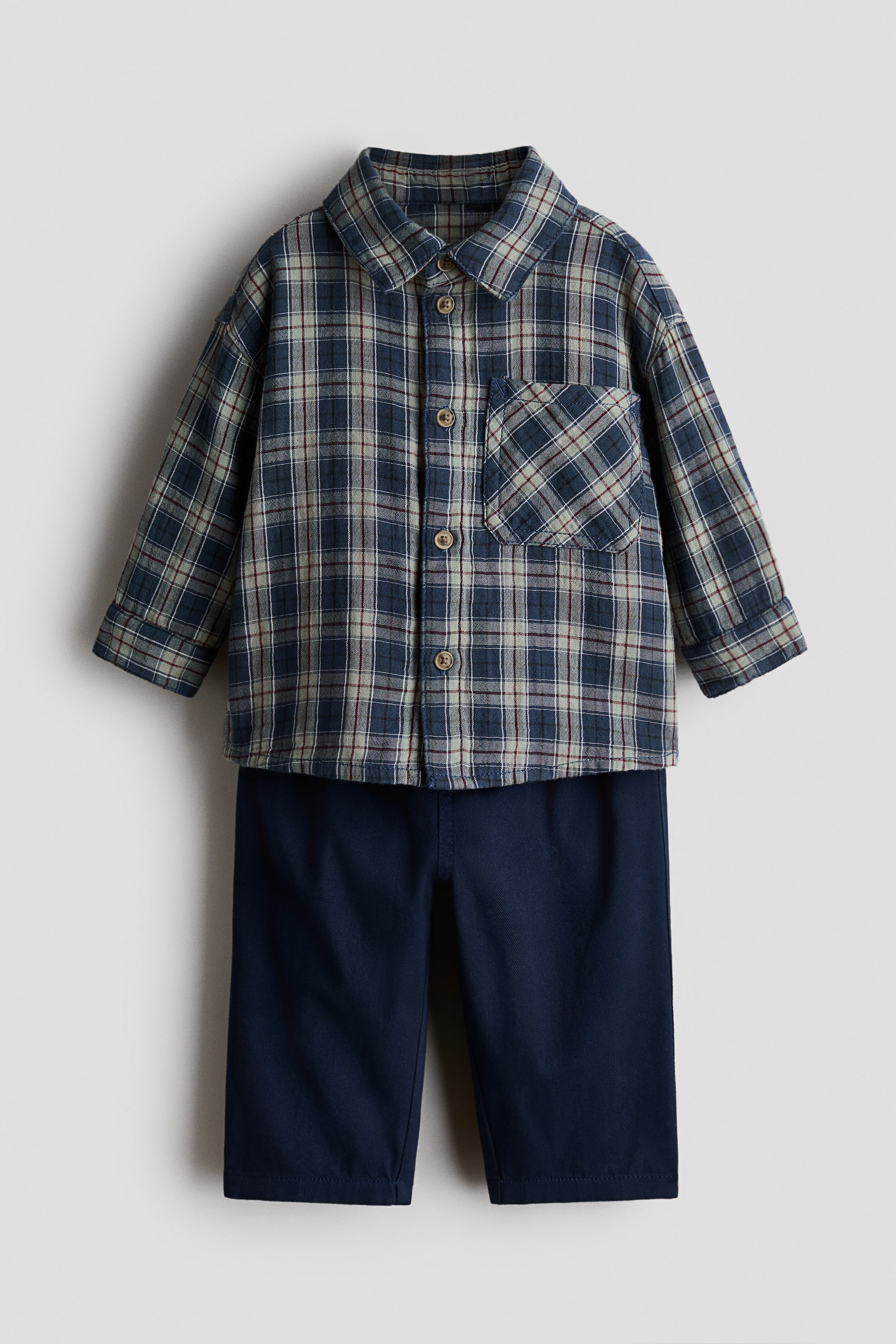 View larger image: A baby's two-piece outfit comprises a long-sleeved plaid shirt and solid navy blue pants, showcased from a front perspective. The shirt features a collar, a full button-front closure with round buttons, and a single patch pocket on the left chest. The plaid pattern highlights shades of blue, grey, and thin red lines.