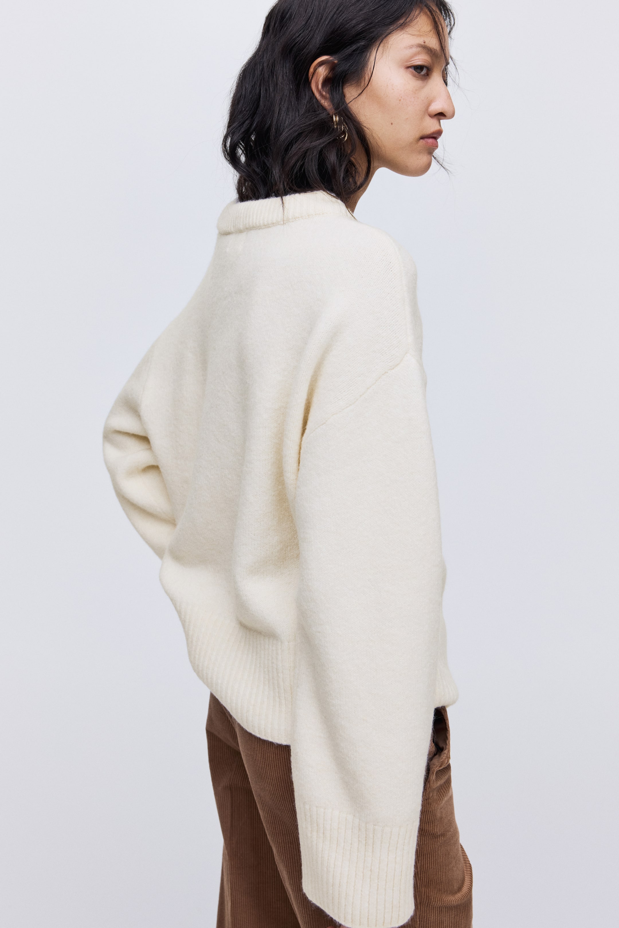 View larger image: An off-white, relaxed-fit sweater with a soft, slightly fuzzy knit texture is being viewed from the back and right side, featuring a high ribbed collar. Its long sleeves end in wide ribbed cuffs, and the ribbed hem falls loosely over brown corduroy pants.