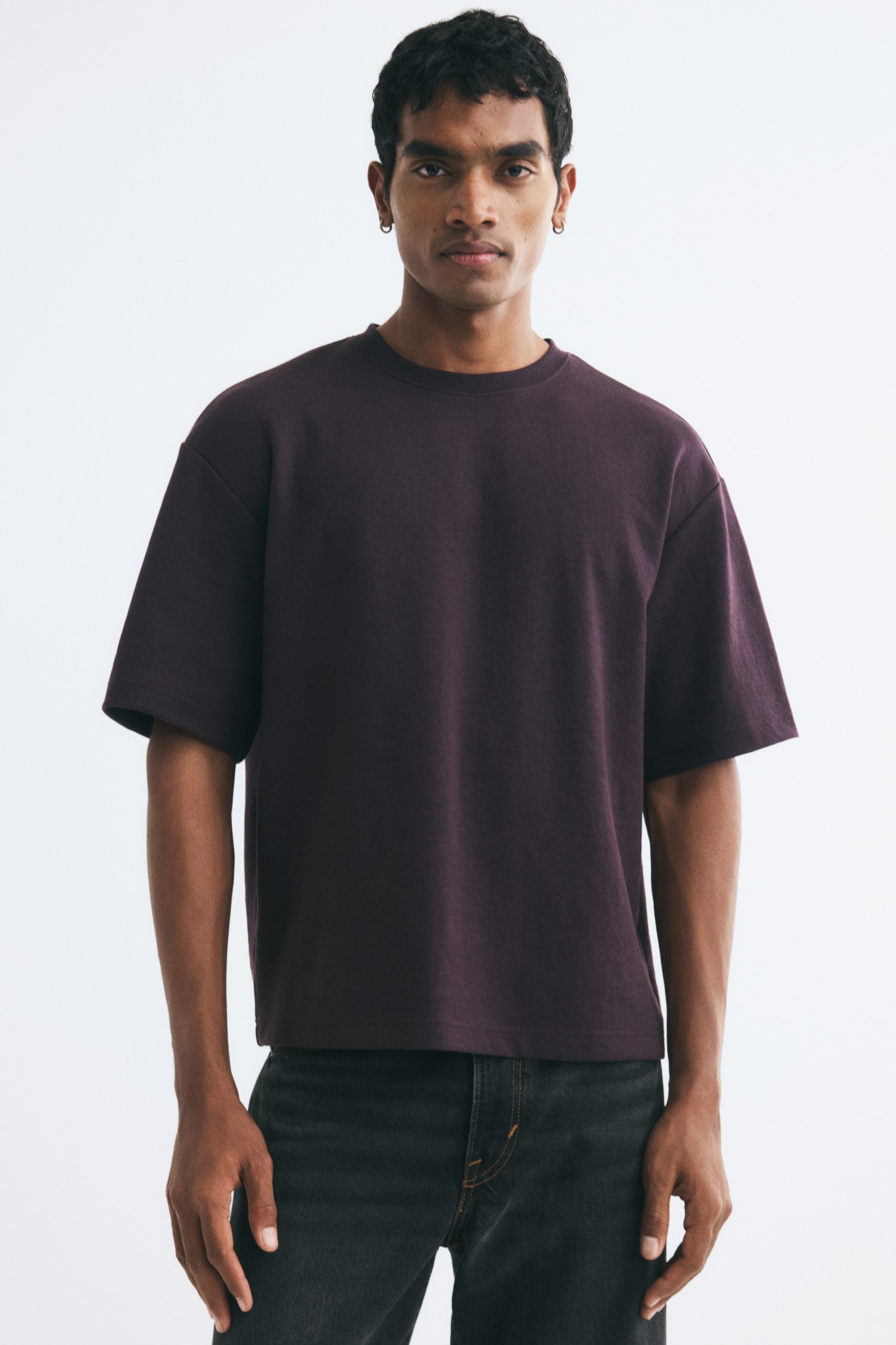 View larger image: A dark purple heavyweight jersey T-shirt is being worn by the model, featuring a relaxed fit, a round rib-trimmed neckline, dropped shoulders, and a straight hem. The T-shirt is paired with dark grey jeans.