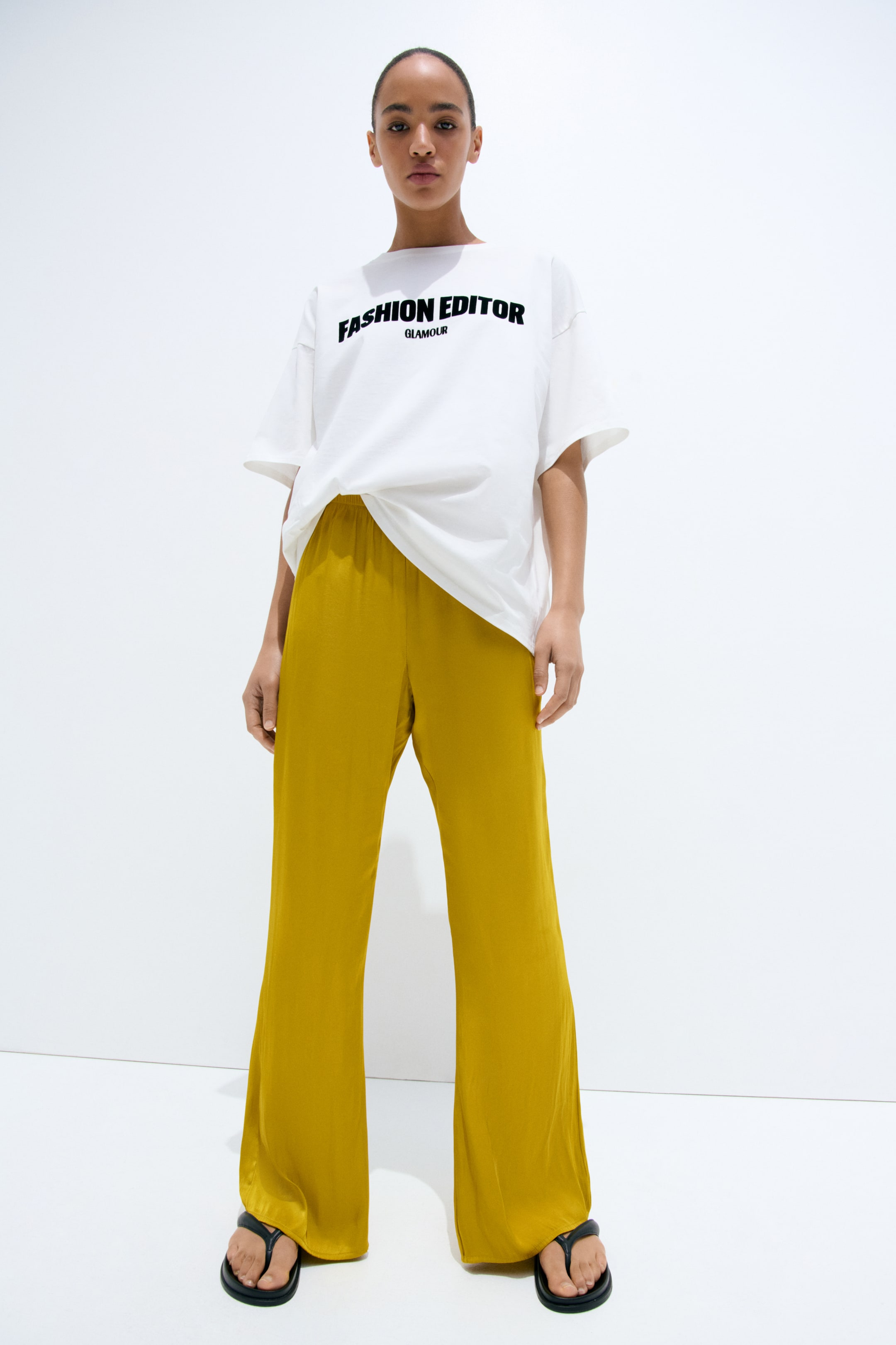 View larger image: The white oversized t-shirt showcases a black "FASHION EDITOR GLAMOUR" graphic on its front. It features a wide crew neck and short, wide sleeves, and is partially tucked at the front.
