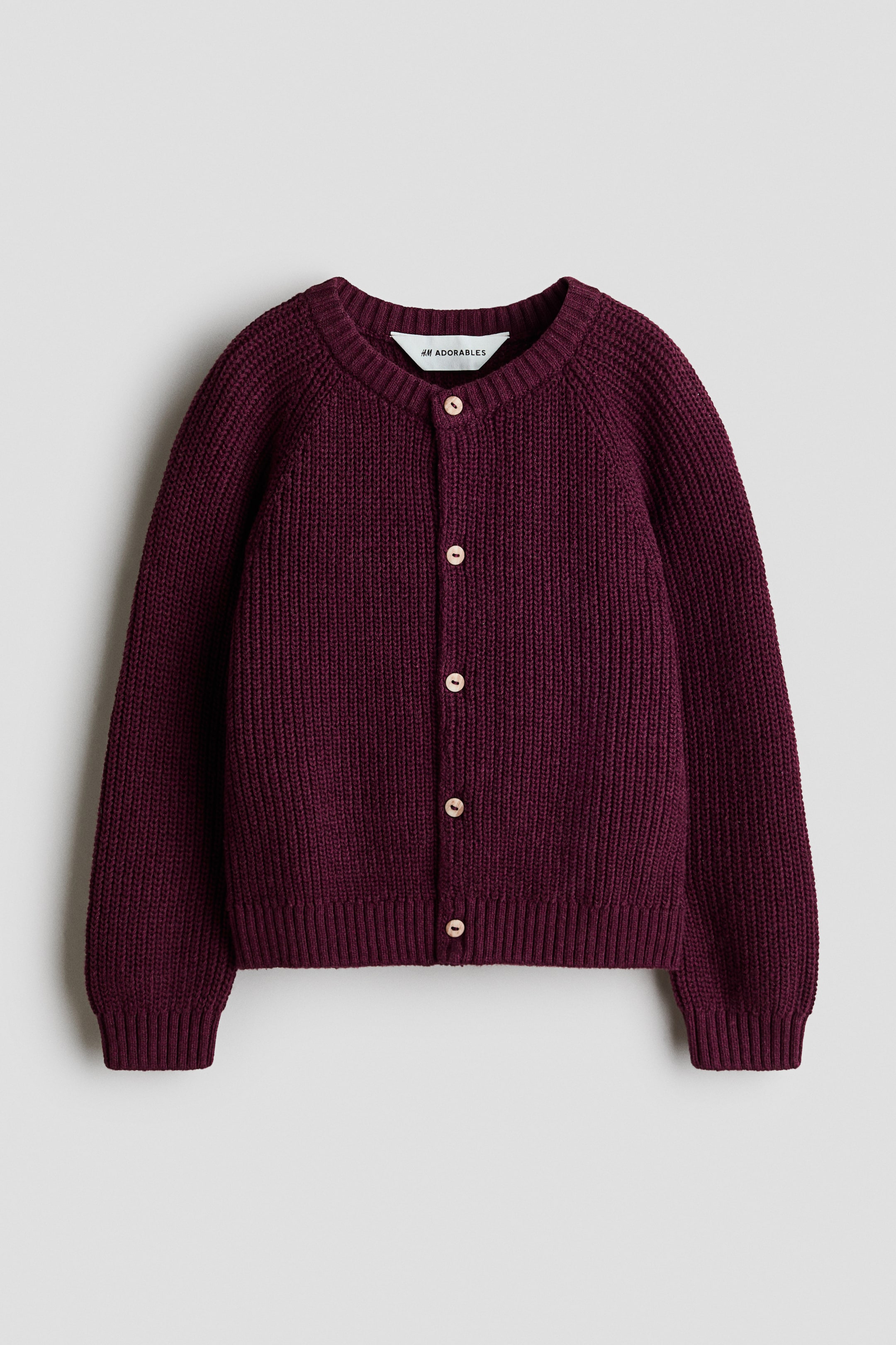 Agrandir l'image: Un cardigan H&M Adorables d'un rouge profond est présenté de face, mettant en avant une texture en maille côtelée. Il est conçu avec un col rond bordé de côtes et de légers boutons circulaires qui descendent sur le devant. Des manches raglan longues sont visibles, avec des poignets et un ourlet côtelés qui complètent le vêtement.