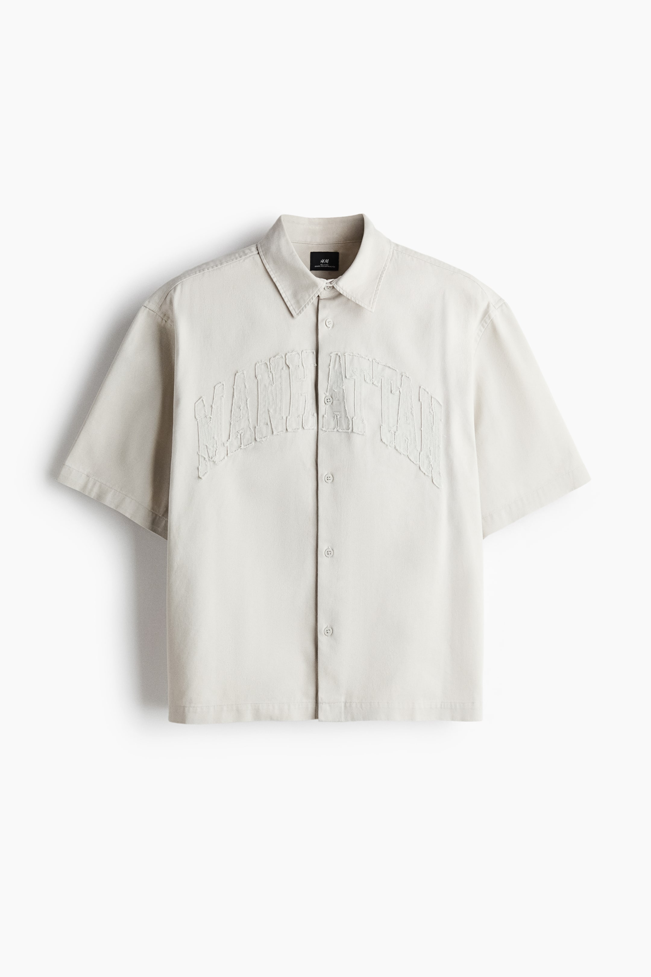 View larger image: A light beige short-sleeve shirt is shown from the front, featuring a spread collar and a button-front closure with matching buttons. The shirt displays the arched word “MANHATTAN” in a distressed, raised texture across the chest.
