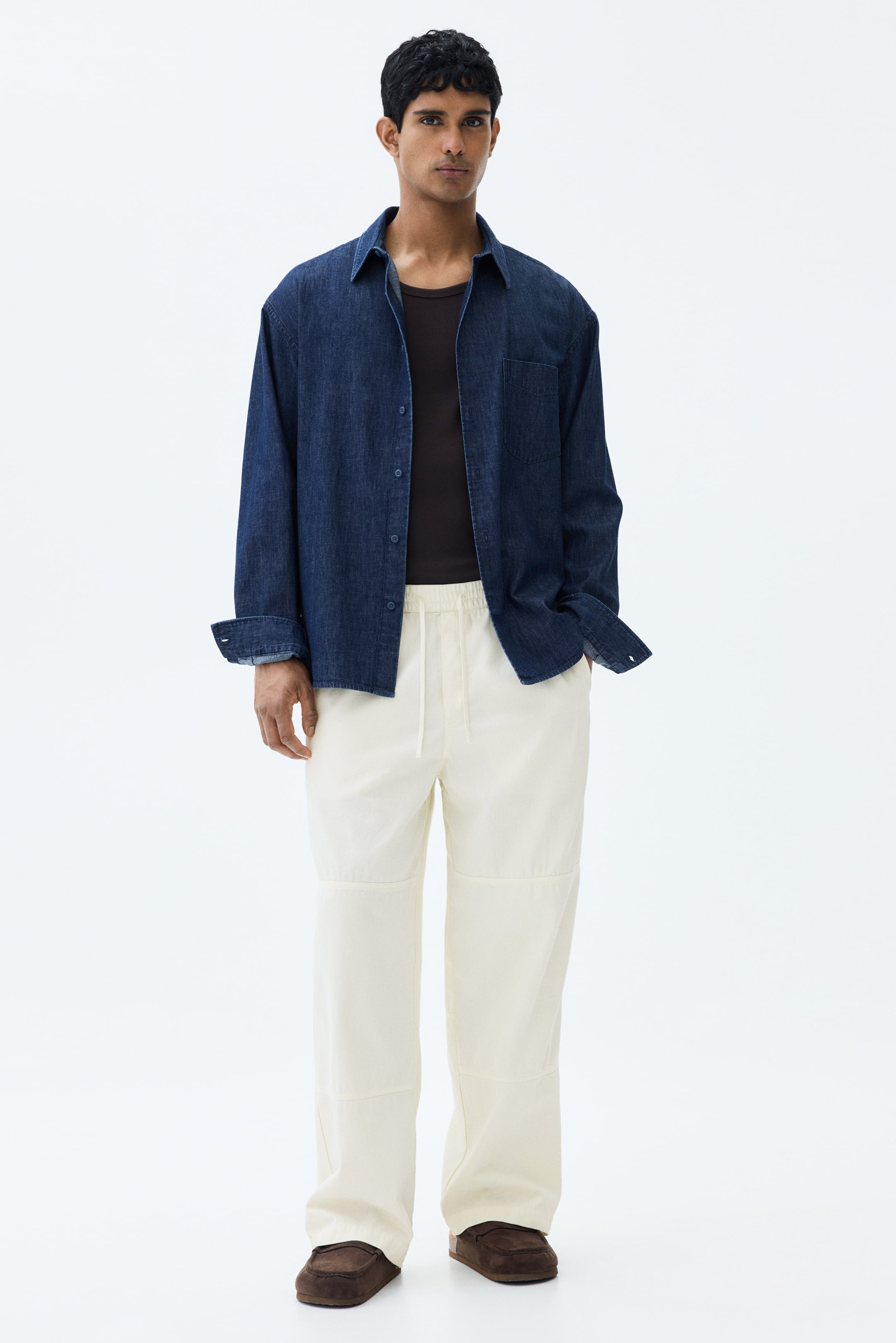 View larger image: The model is showcasing light cream trousers with a relaxed fit. These trousers are designed with a visible drawstring and elasticated waist, diagonal side pockets, and panel detailing on the legs. The model's left hand is casually placed in one of the side pockets.