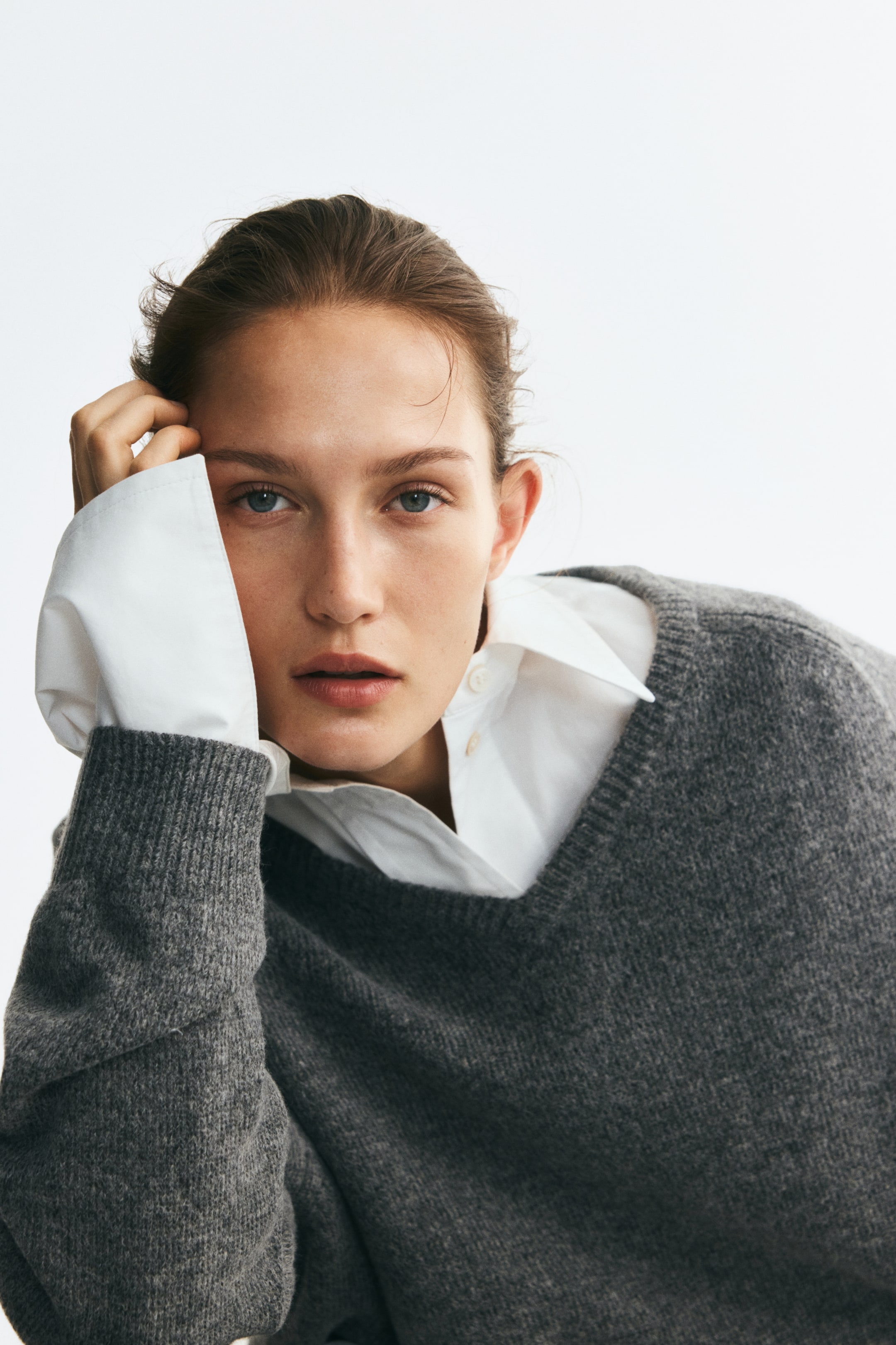 Agrandir l'image: Un pull gris en maille douce à col en V est porté par le mannequin par-dessus une chemise blanche à col. Le pull présente un col bordé de côtes et des poignets côtelés, avec une manche reposant contre la tête du mannequin pendant la pose.