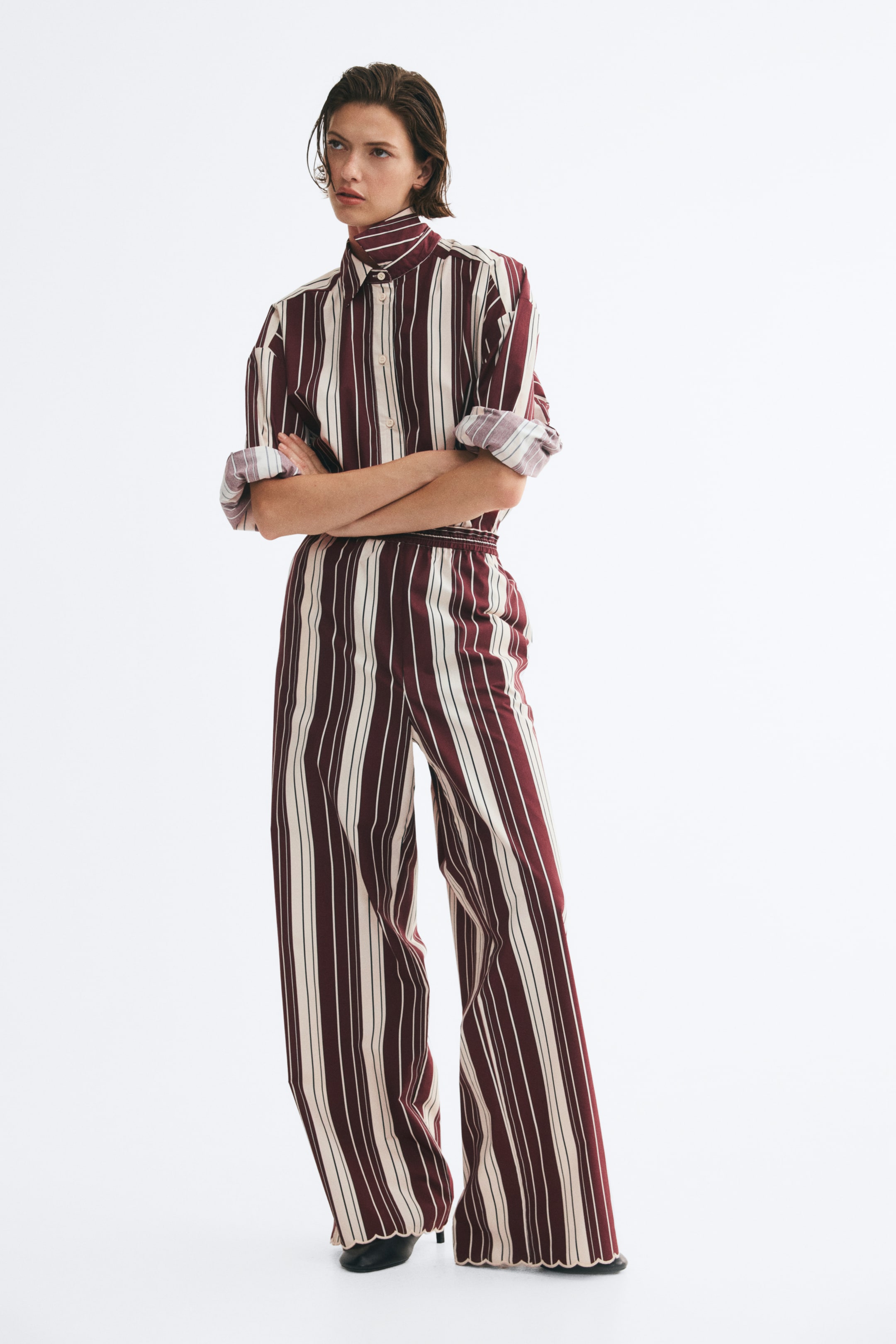 View larger image: Loose-fit trousers in a burgundy and cream vertical stripe pattern are worn by the model, featuring a high, elasticated waist and straight legs with scalloped hems. The trousers are styled with a matching tucked-in, striped long-sleeve shirt.