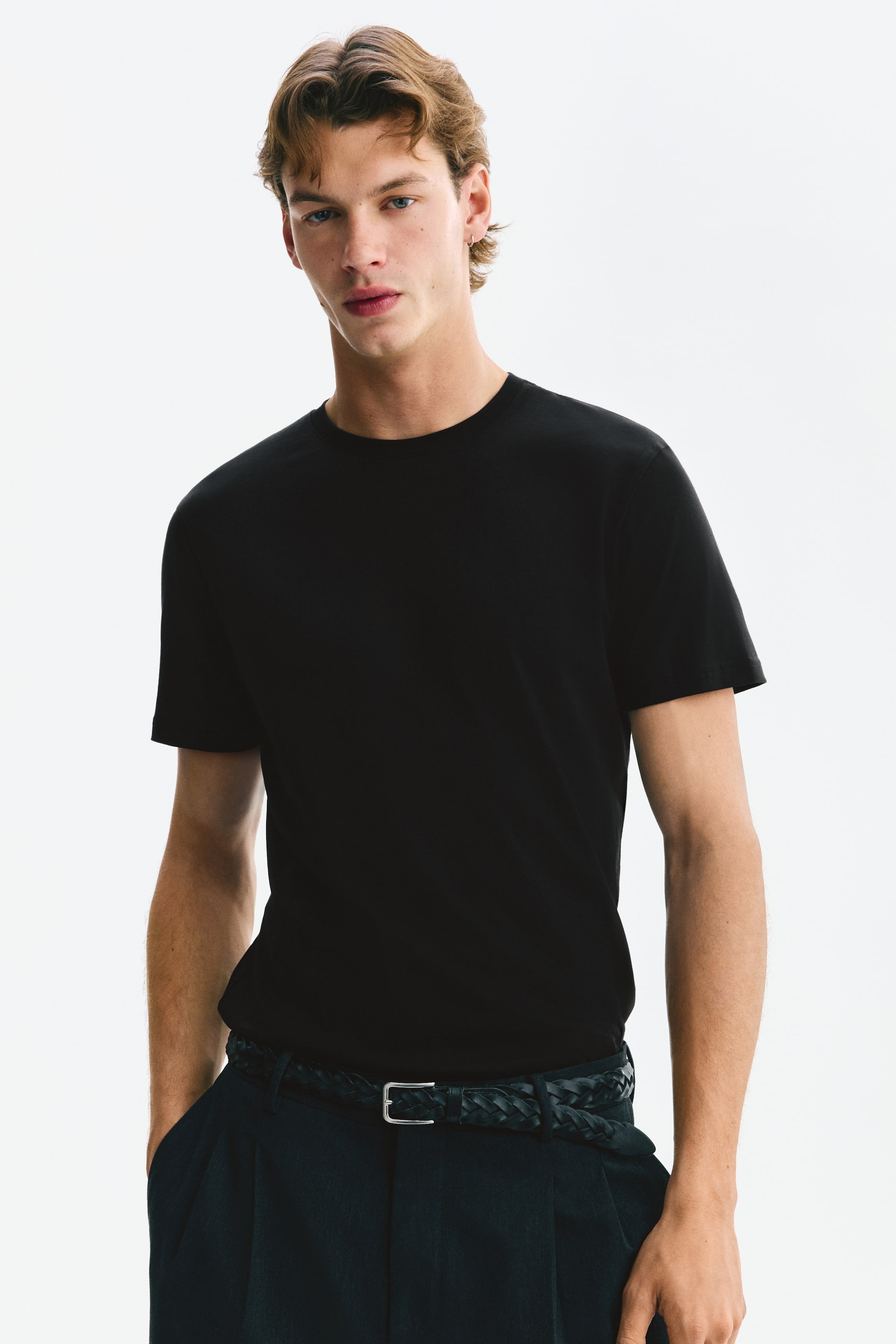 Visa större bild: En svart slim-fit t-shirt med rund halsringning och finribbad kant samt korta ärmar bärs av modellen. T-shirten är instoppad i mörka byxor med ett synligt flätat bälte, och modellen står med ena handen i en ficka.
