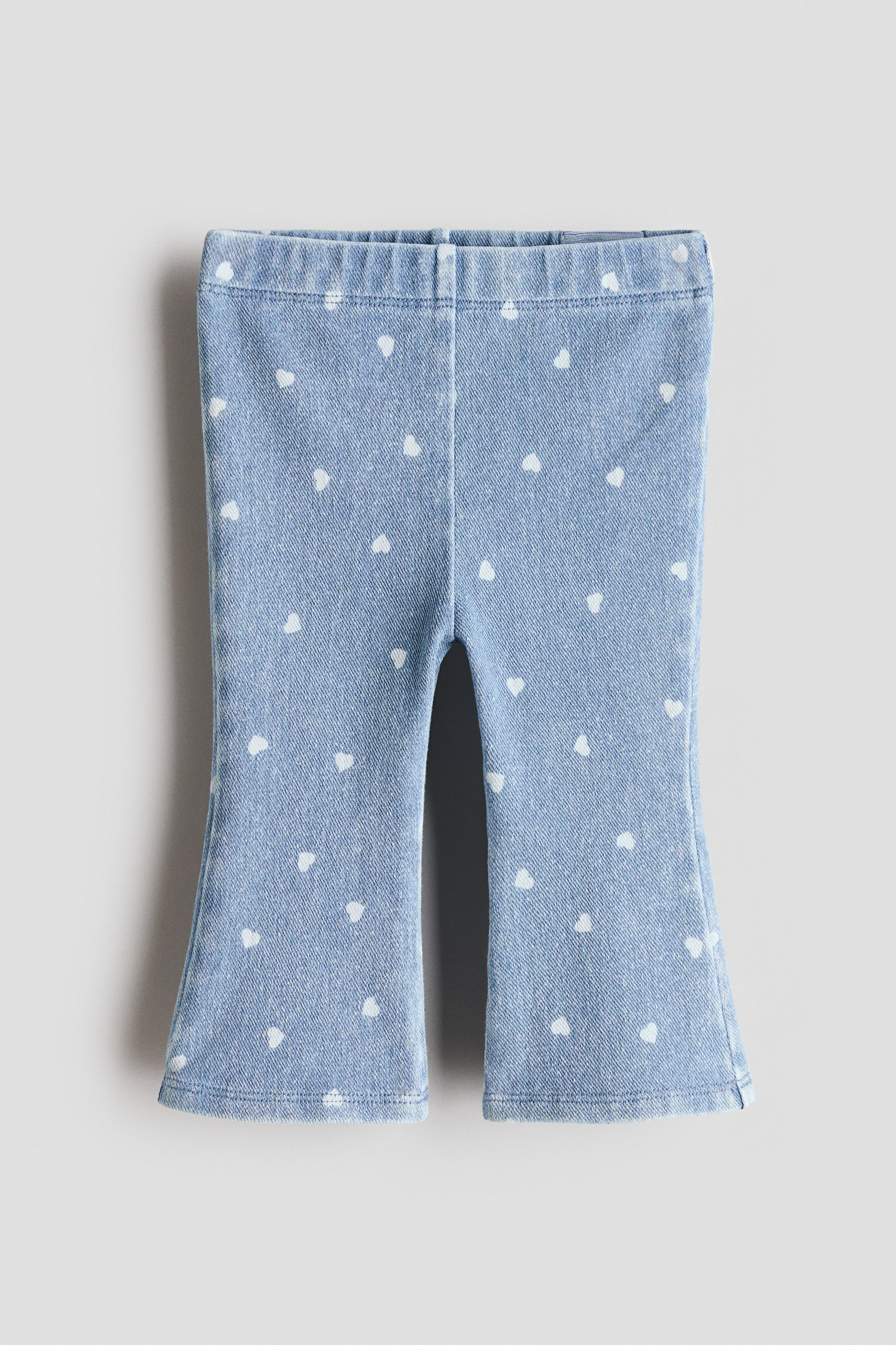 Ampliar la imagen: Los pantalones para bebé están presentados de frente, con una tela de imitación denim azul claro con un estampado de corazones blancos pequeños por todas partes, una cintura elástica y piernas que se ensanchan en la parte inferior.