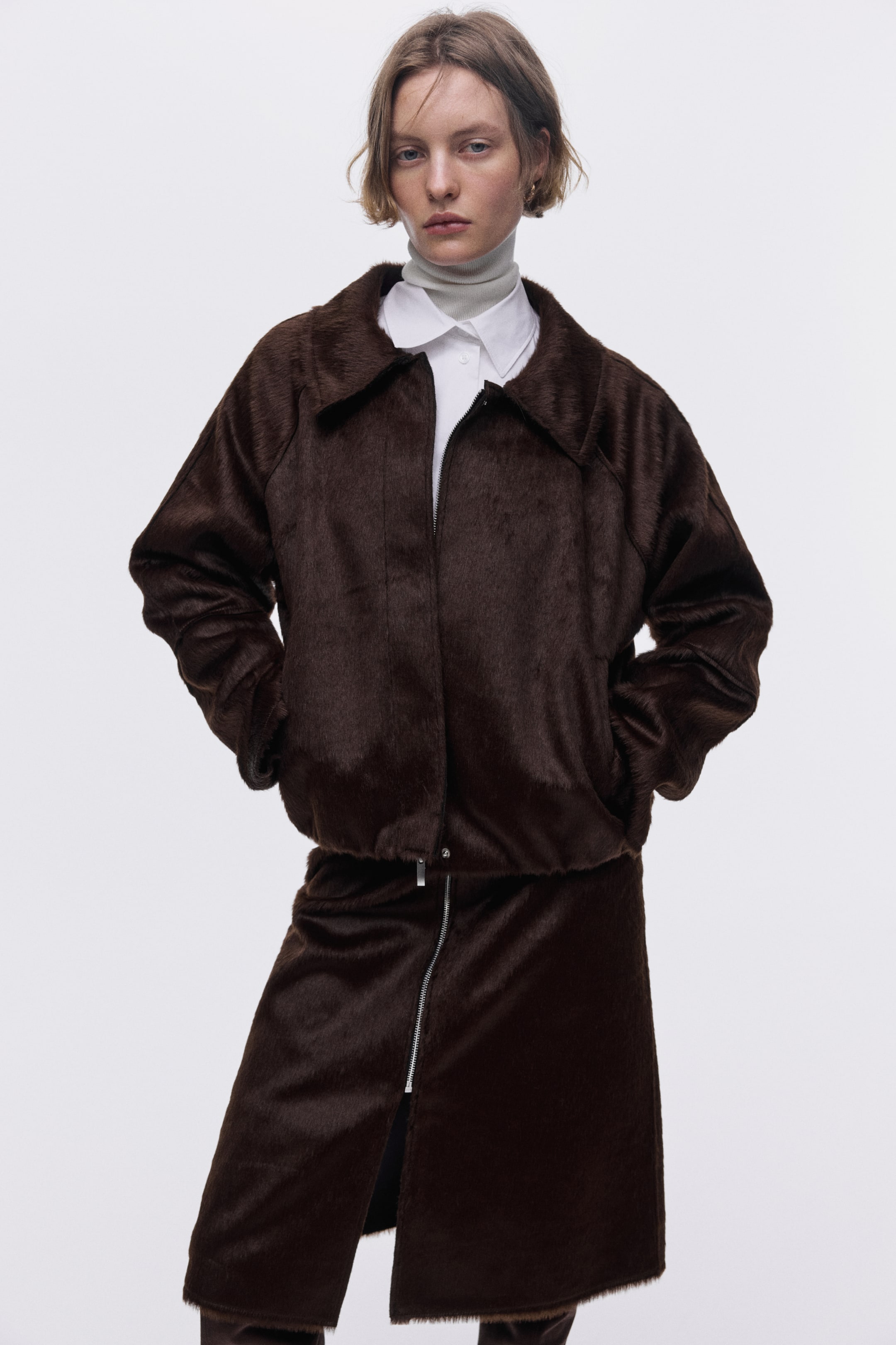 拡大画像を表示: 濃い茶色のショートパイルのファーのようなジャケットとそれに合うミディスカートが特徴です。ジャケットは、尖った襟、前面のジッパー開閉、サイドポケットがあり、白い襟シャツとグレータートルネックの上にオープンに着用されています。スカートは、ハイスリットを作る前面のジッパーを備えています。