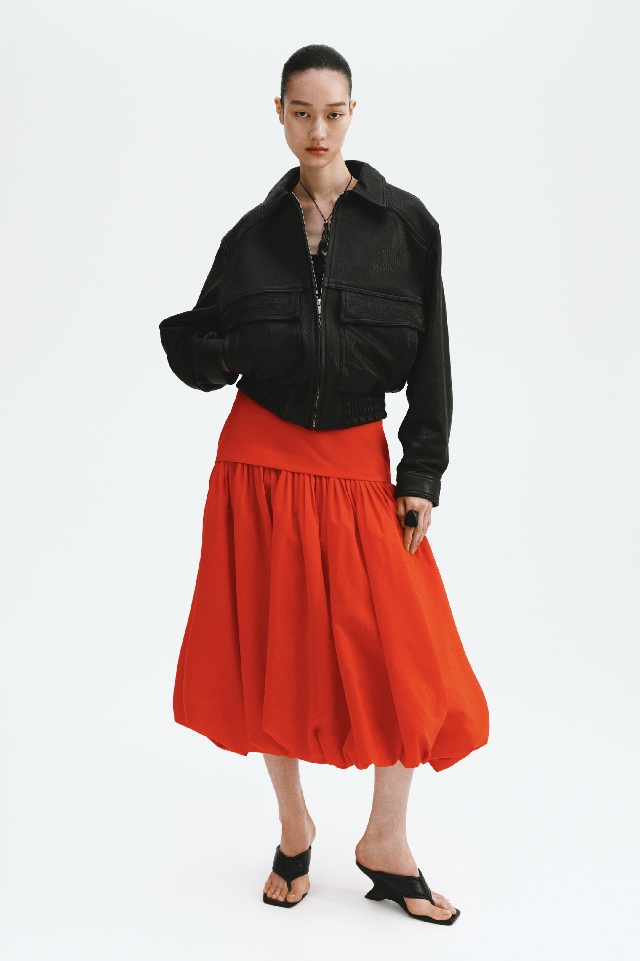 Grösseres Bild ansehen: Eine schwarze, strukturierte Bomberjacke mit einem Umlegekragen, einem Frontreißverschluss und zwei Pattentaschen mit horizontalen Reißverschlusstaschen, die über einem elastischen Saum liegen. Ein lebendiger roter voluminöser Midirock ergänzt die Jacke und präsentiert einen breiten, flachen Bund sowie eine geraffte Blasensilhouette.