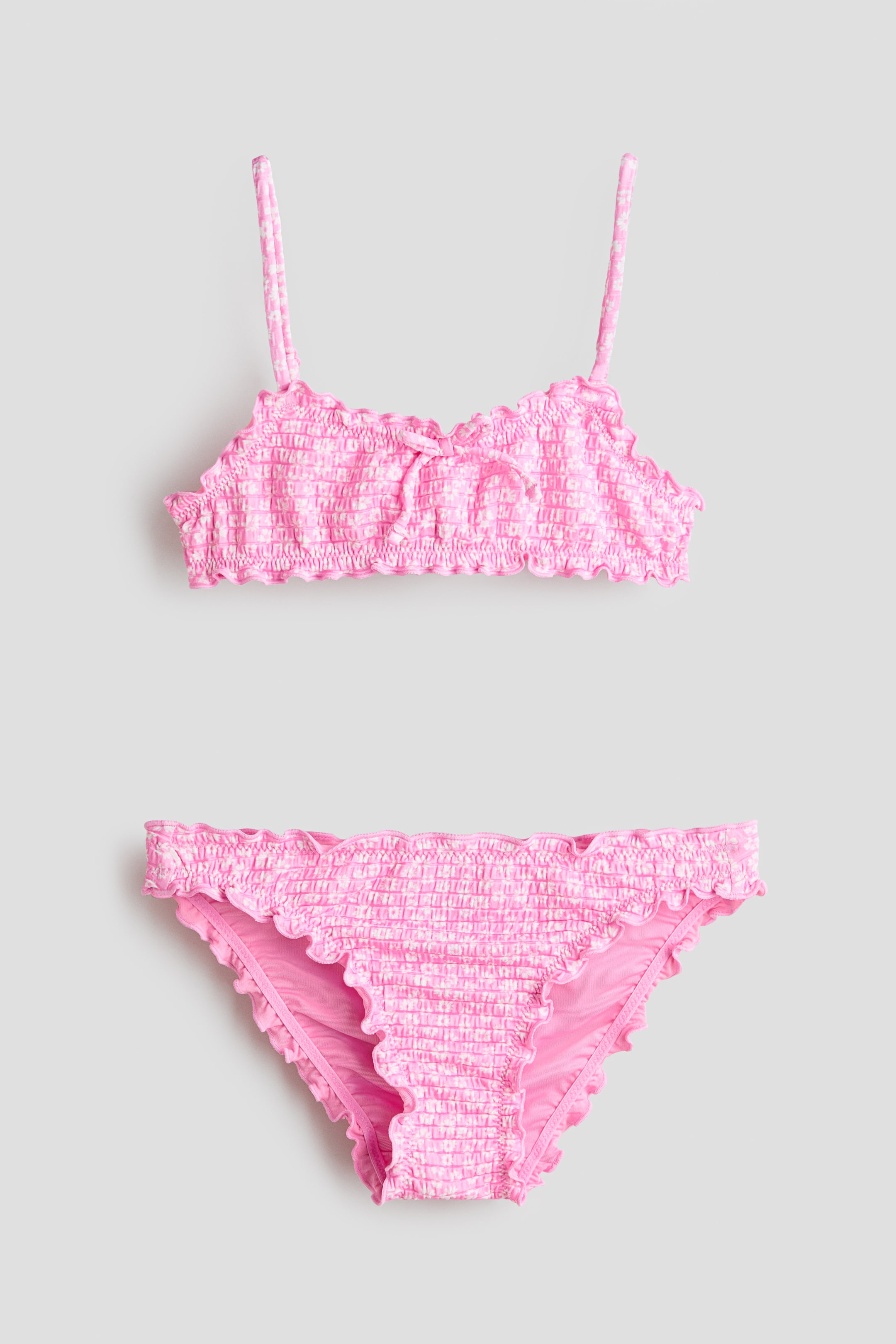 View larger image: A fully lined pink and white gingham bikini set showcases a smocked texture throughout. The bikini top features narrow, adjustable shoulder straps and a sewn-in bow at the front, while both the top and matching bottoms display ruffled, overlocked edges.
