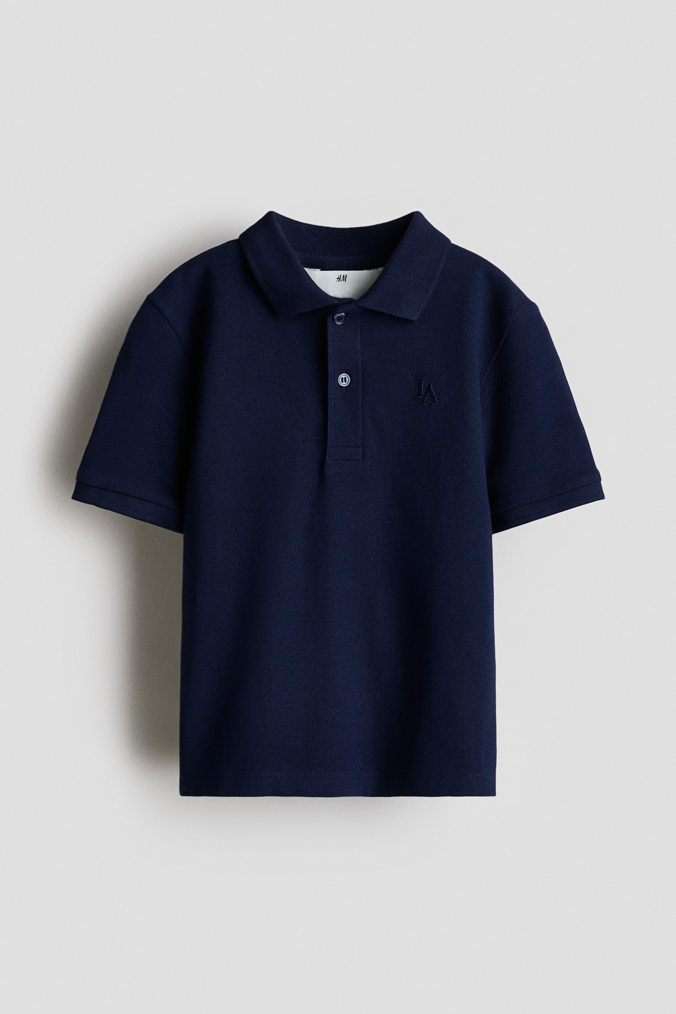 View larger image: A navy-blue polo shirt shown from the front, featuring a piqué knit texture, a classic pointed collar and a two-button placket. It has short sleeves with cuffed hems and a subtle 'LA' embroidery on the left chest.