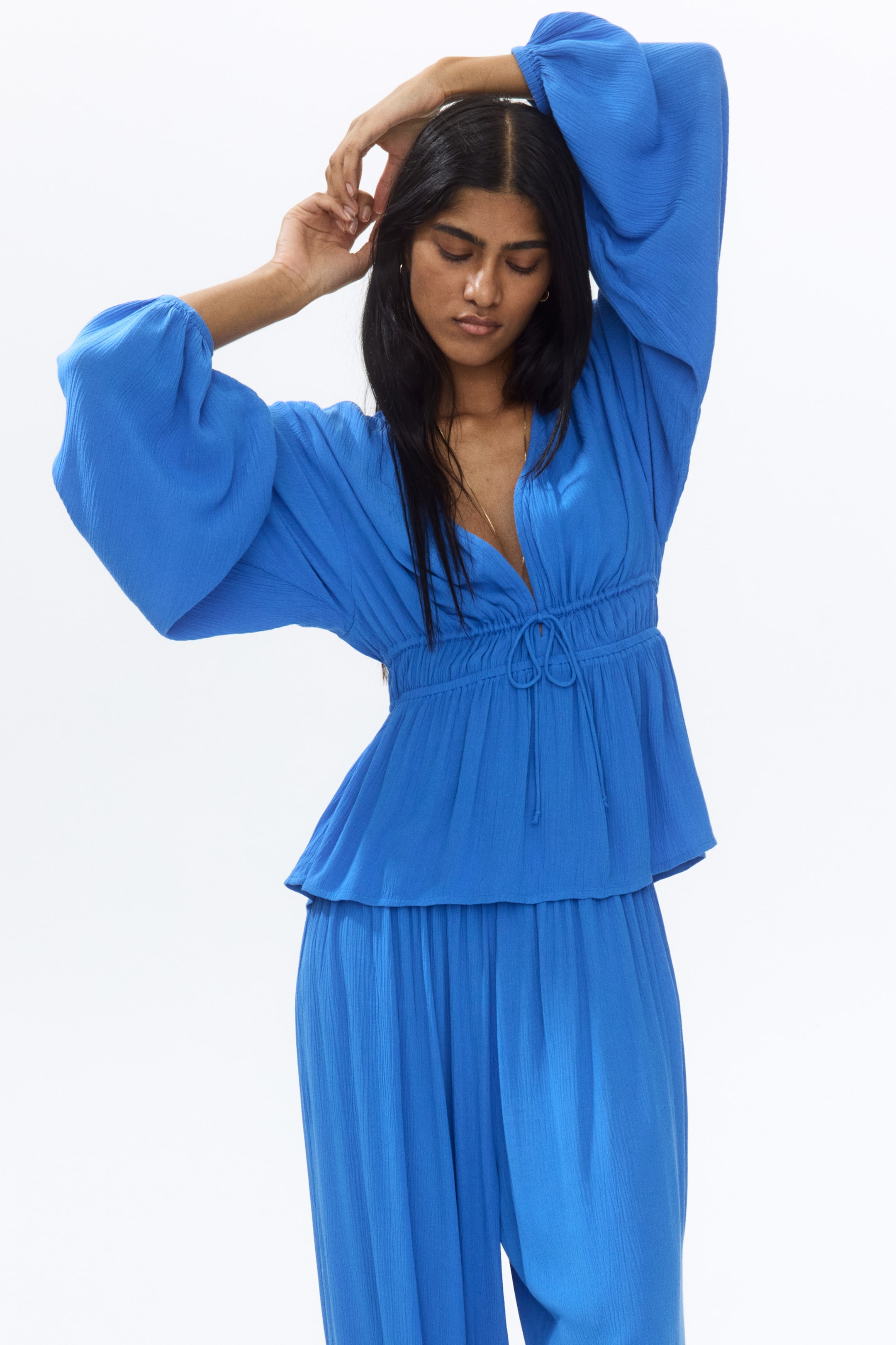View larger image: The model is donning a blue blouse crafted from a crinkled viscose weave, showcasing a deep V-shaped neckline. It features long balloon sleeves with narrow elasticated cuffs and a slender drawstring at the waist, which is elasticated to accentuate the silhouette. The model is raising her arms to highlight the blouse, paired with matching blue trousers.
