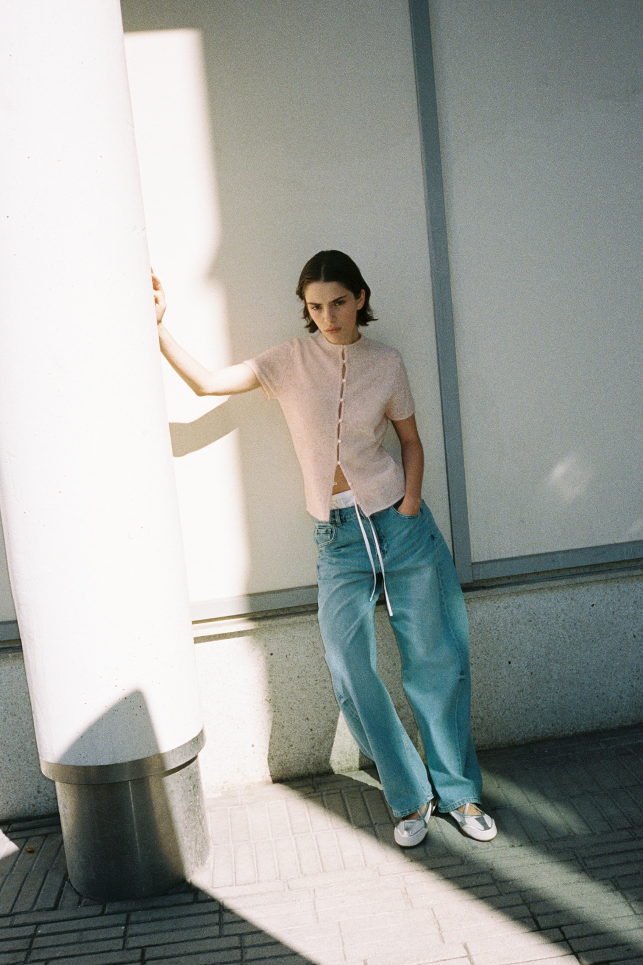 Voir une image plus grande: Un cardigan en tricot fin à manches courtes, rose pâle, doté d’un col rond et d’une fermeture à boutons et brides à l’avant, est porté par le mannequin. Le cardigan ajusté est porté avec un jean bleu à jambes larges et stylisé avec les boutons du bas laissés ouverts.