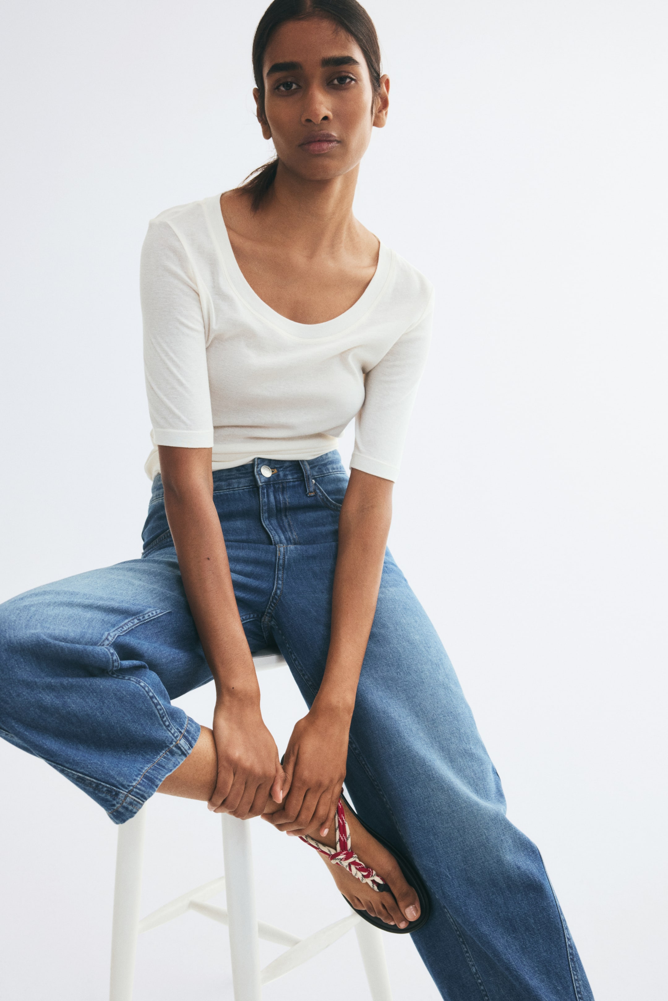 View larger image: Blue denim jeans featuring a regular waist and curved, voluminous legs are being worn by the model, who is sitting on a white stool. The medium-wash jeans are styled with a white scoop-neck top and flat sandals, highlighting their relaxed silhouette.