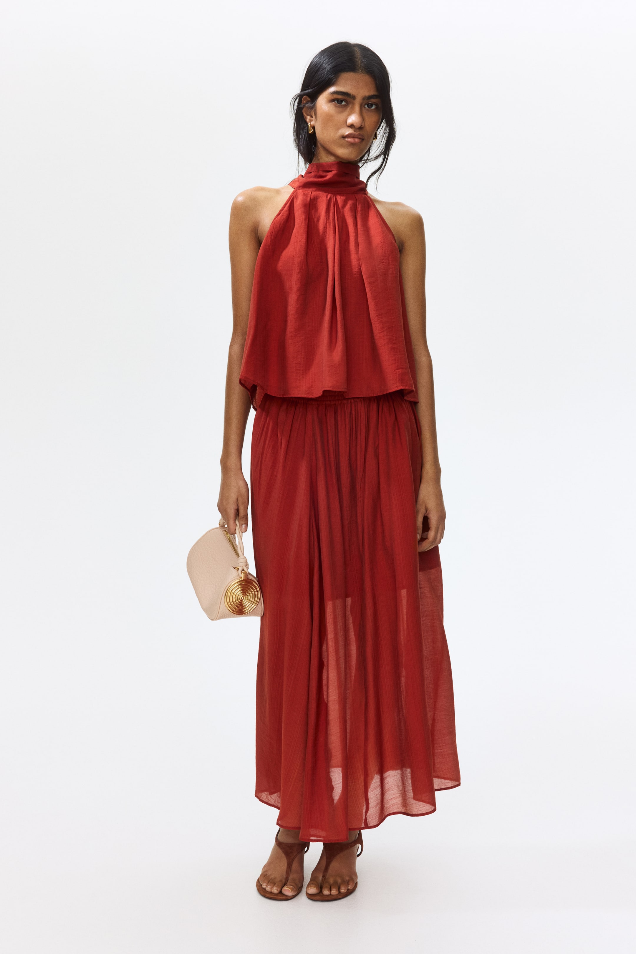 View larger image: A rust-coloured two-piece outfit showcases a graceful halter-neck top with gathers around the high neckline, paired with a matching floor-length skirt featuring a gathered elasticated waist. The ensemble is accessorised with a light beige structured handbag, adorned with a gold spiral detail, and complemented by brown flat sandals.
