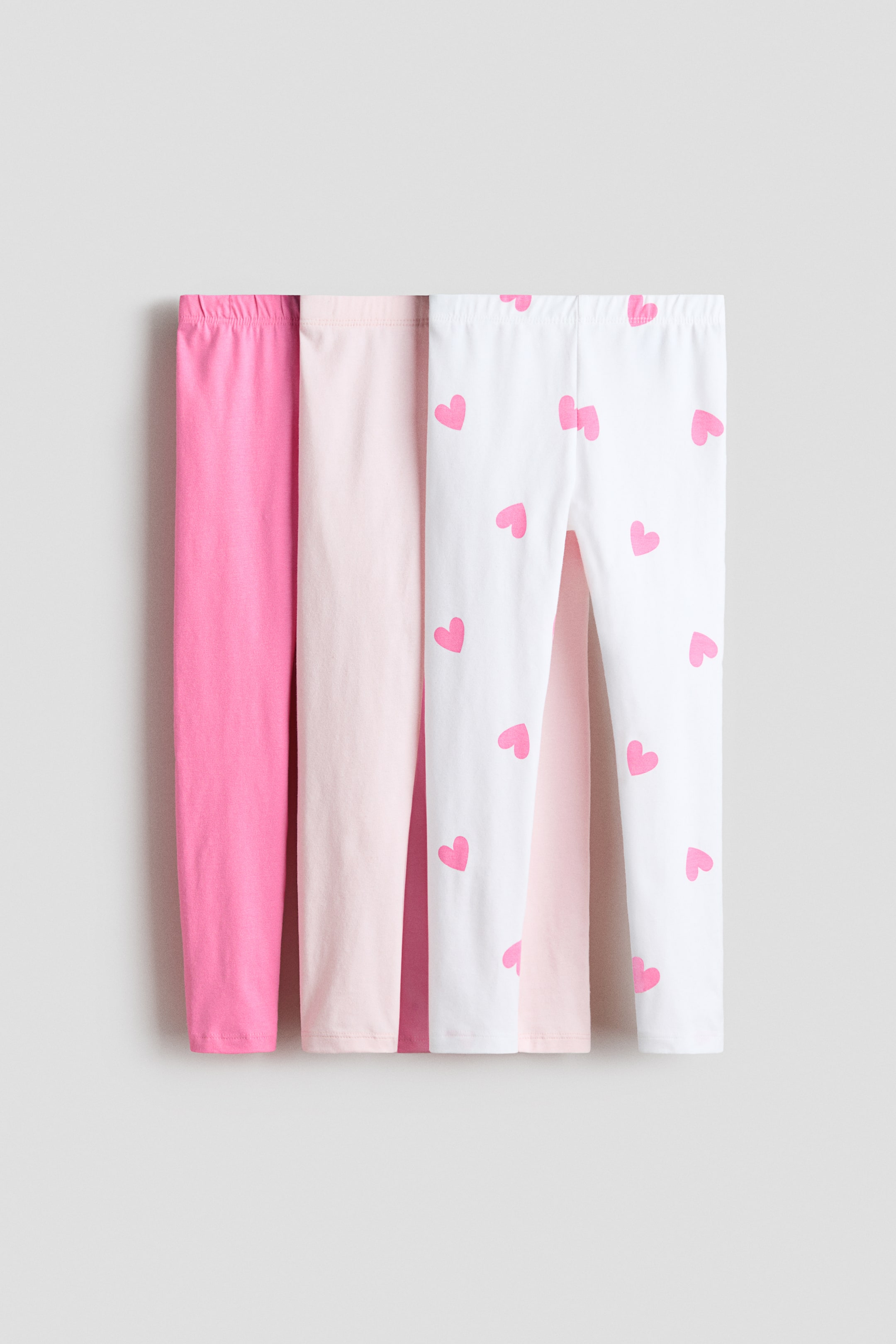 View larger image: Three pairs of leggings are displayed, showing their front. The leftmost pair is a solid bright pink, the middle pair is a solid light pink, and the rightmost pair is white with scattered pink heart patterns. All leggings feature elasticated waistbands.