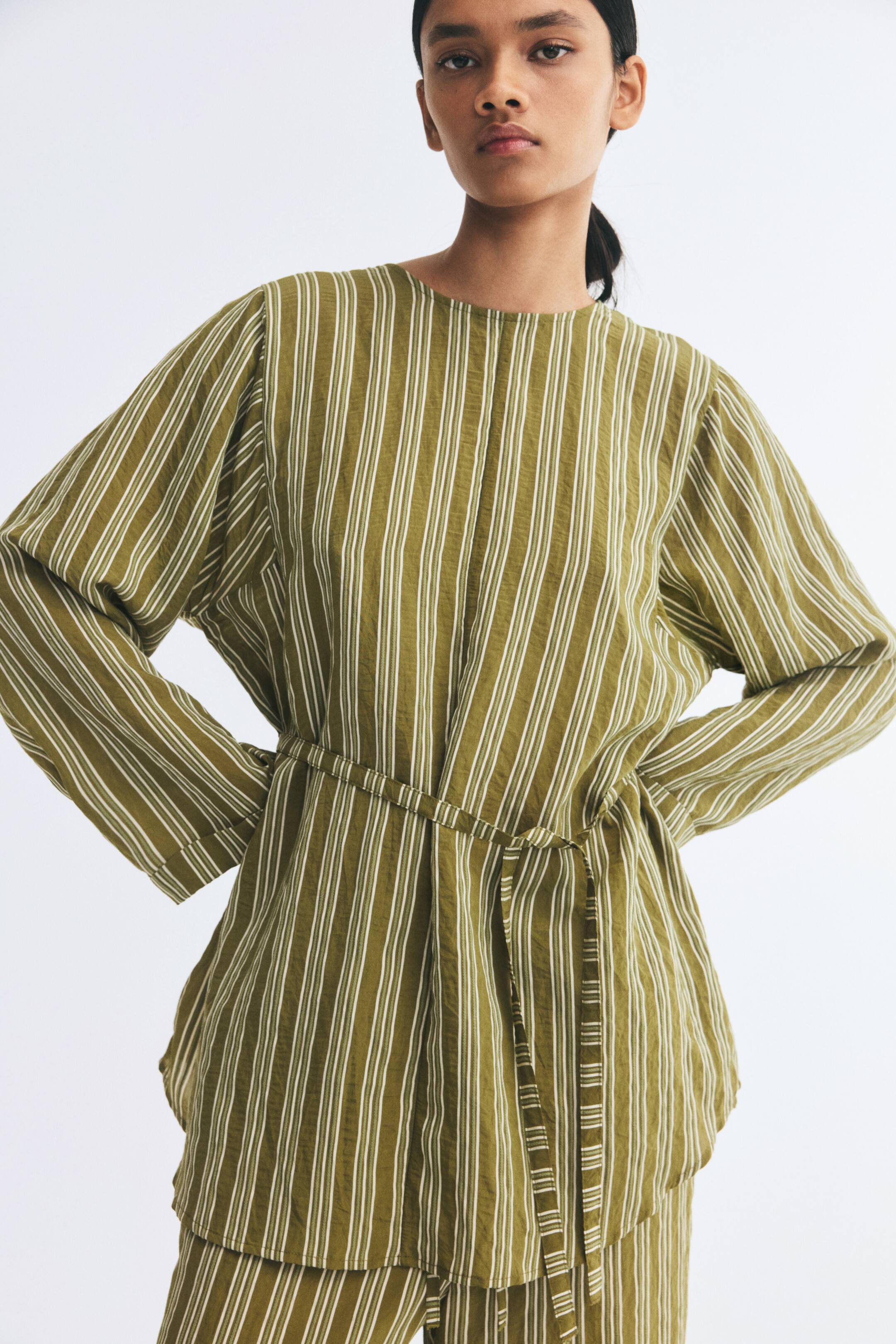 View larger image: An oversized blouse in a green and white striped textured weave is being worn by the model. The blouse features a round neckline, long sleeves with turn-up cuffs, and a narrow tie belt at the waist, paired with matching striped trousers.