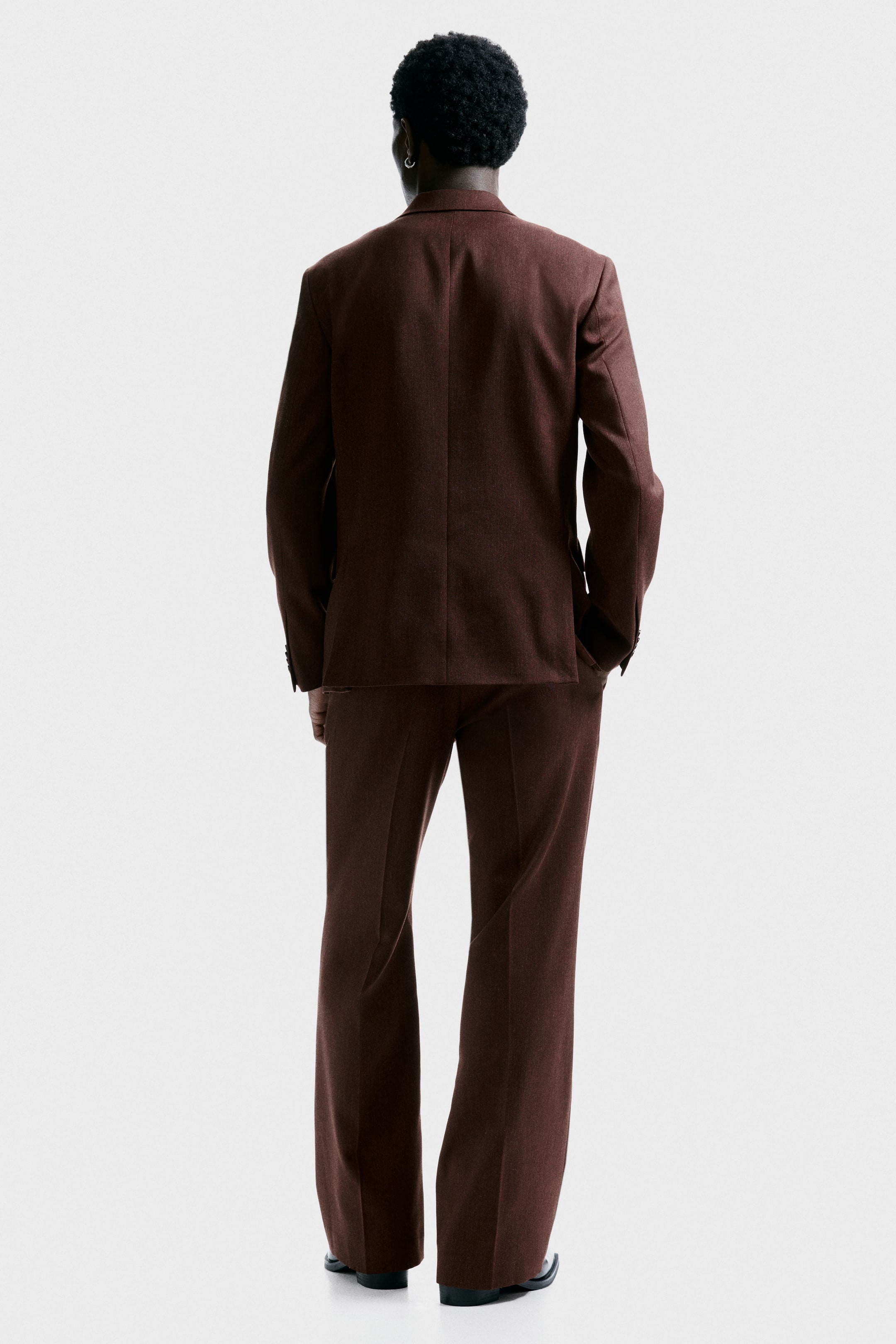 拡大画像を表示: 暗い茶色のスーツは、微細な織り模様を特徴としており、後ろから紹介されています。ジャケットは、ベンツのない滑らかな背中を見せており、合わせたパンツはフレアレッグを示しています。