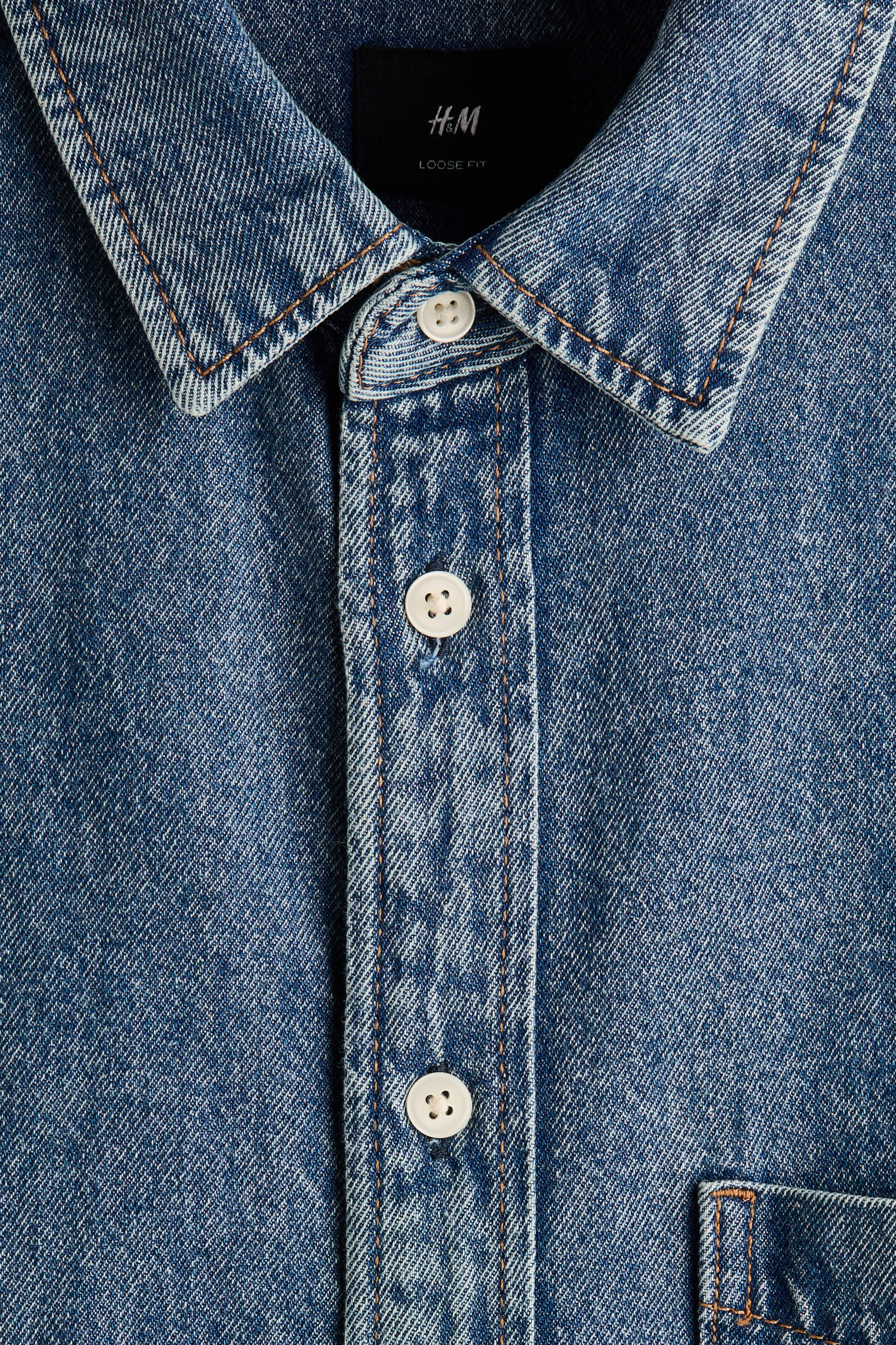 View larger image: A blue denim shirt is presented front-on, featuring a spread collar and a central button placket with white four-hole buttons. Orange contrast stitching is accentuating the collar, placket, and a visible chest pocket, with a black H&M tag showing "LOOSE FIT" at the neck.