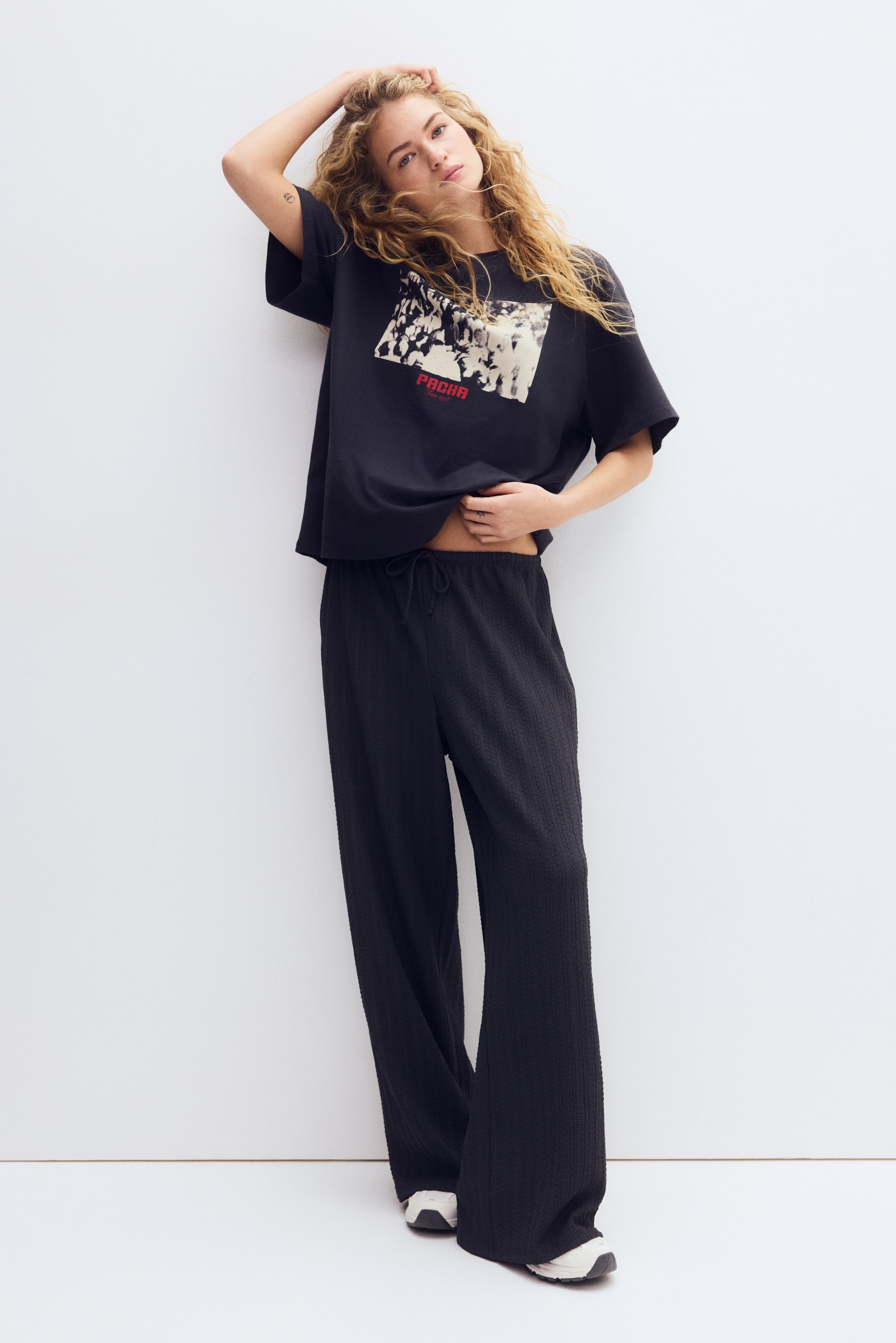 查看大圖: 一件黑色寬鬆T恤，展示一幅長方形黑白群眾圖案，並以紅色「PACHA」字樣點綴。黑色闊腿褲帶有細微的紋理質感與明顯的抽繩腰身，寬鬆垂至腳踝。
