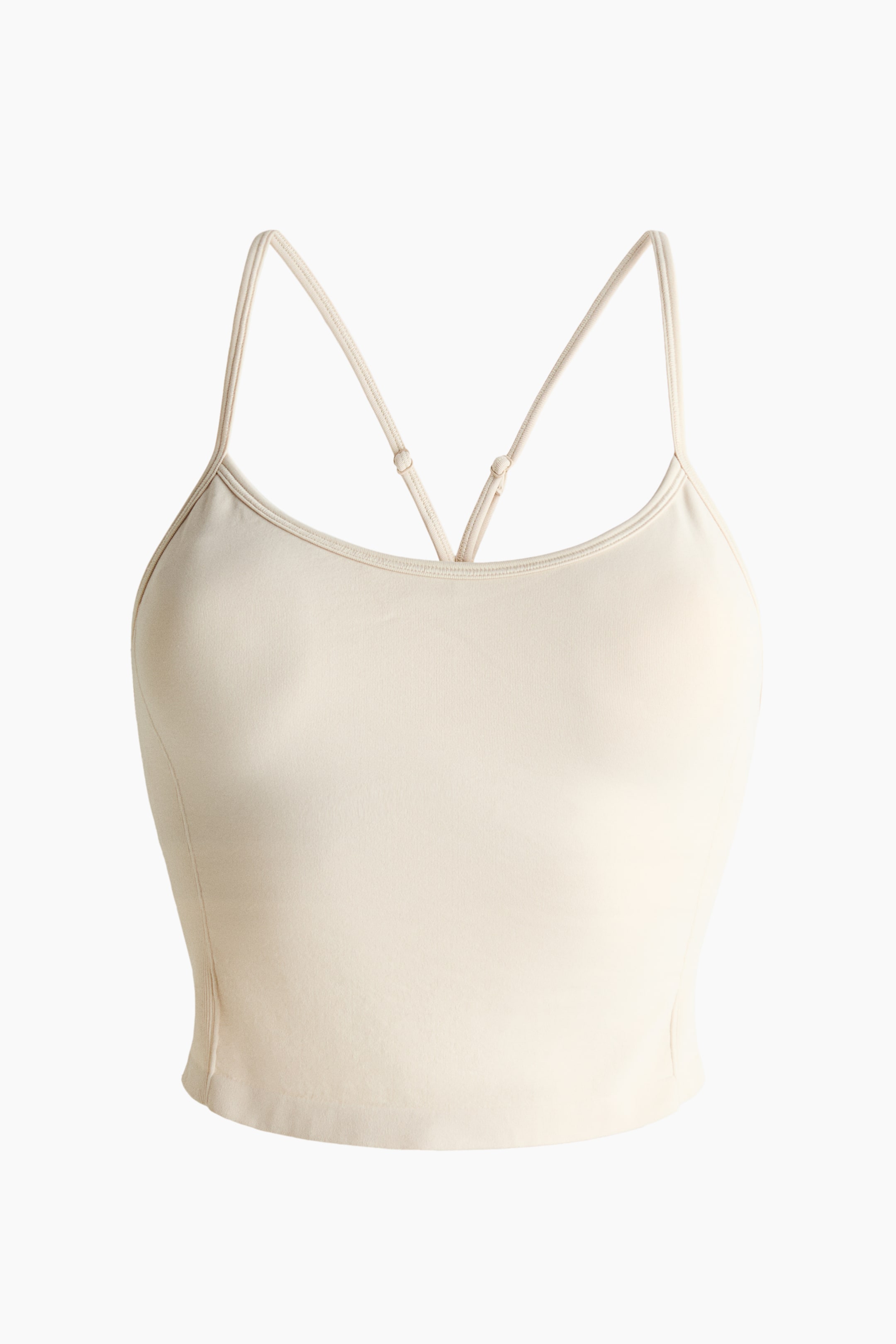 Grotere afbeelding bekijken: Een naadloze sporttop toont zijn vooraanzicht, met een lichte crèmekleur en een ronde halslijn. Het is ontworpen met dunne, verstelbare spaghettibandjes die een kruislingse vorm aan de achterkant vormen, en een kortere lengte.