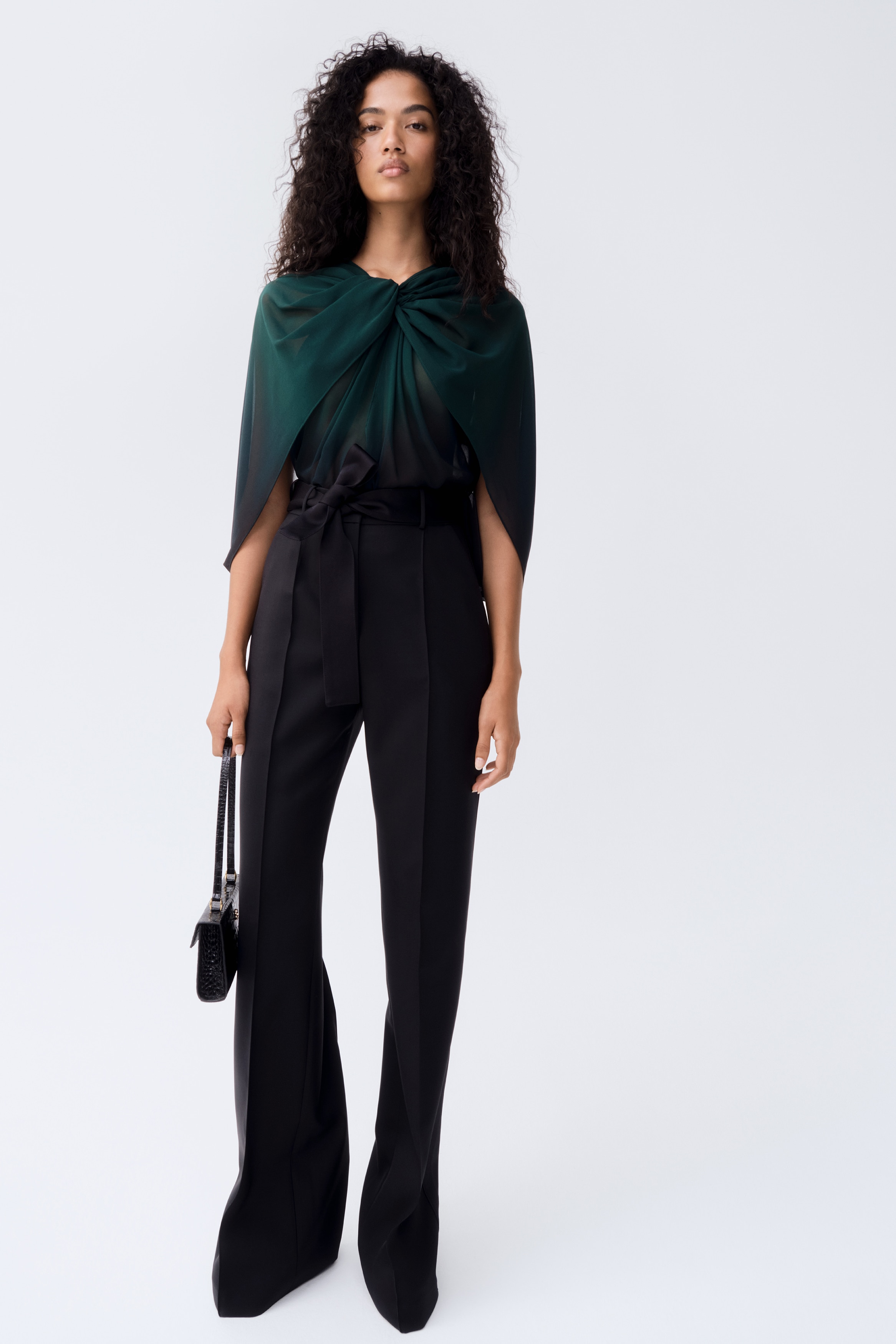 Deze zwarte wollen pantalon met strikdetail wordt frontaal getoond en heeft een hoge taille met een bijpassende stoffen riem die aan de voorkant is geknoopt. Het ontwerp laat rechte, wijde pijpen zien met scherpe, geperste vouwen die over het midden van elke pijp lopen.