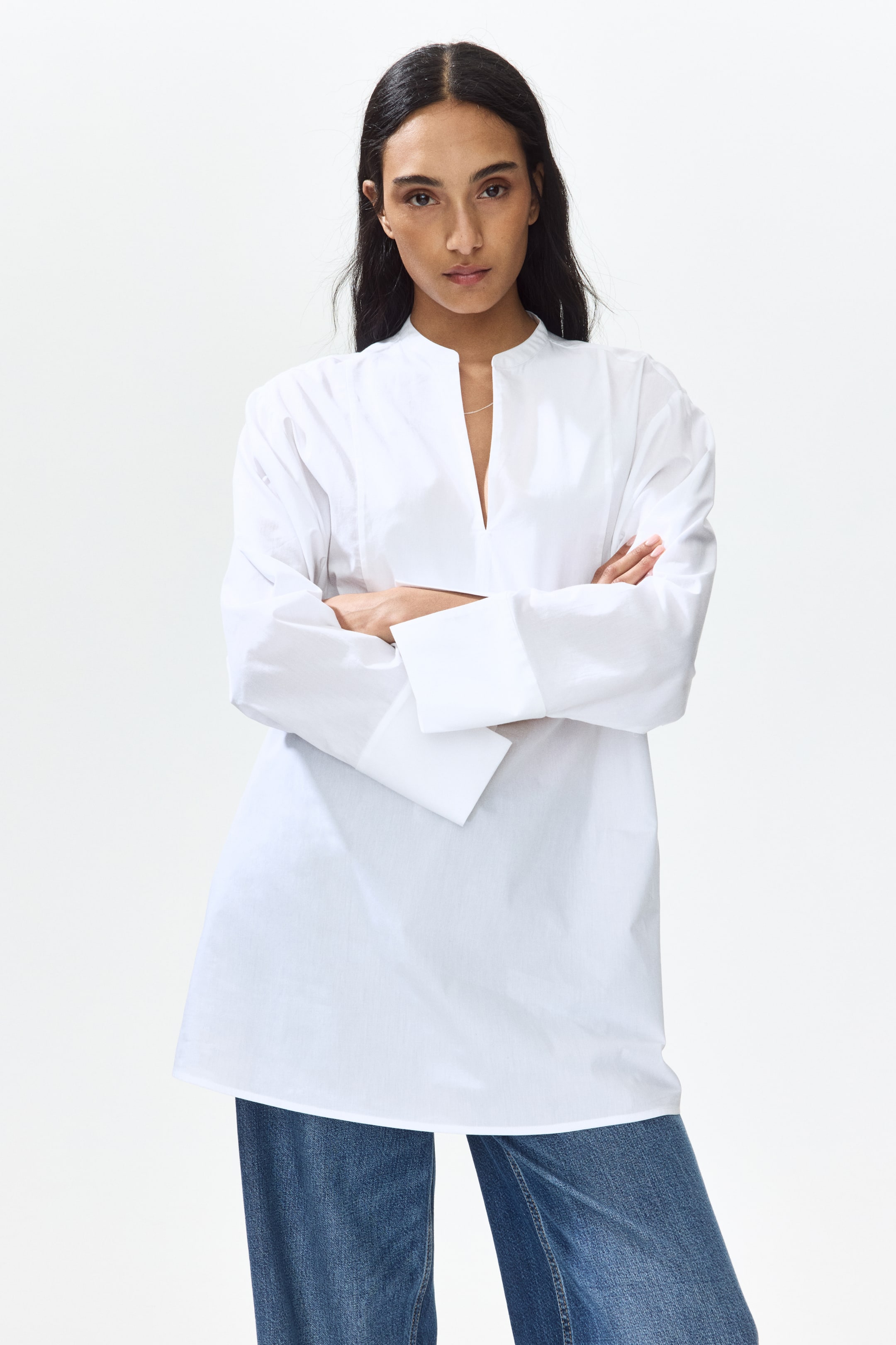 View larger image: A white tunic shirt showcases a band collar with a V-neck opening and long sleeves featuring wide, folded-back cuffs. It offers a relaxed, elongated silhouette that extends beyond the hips.