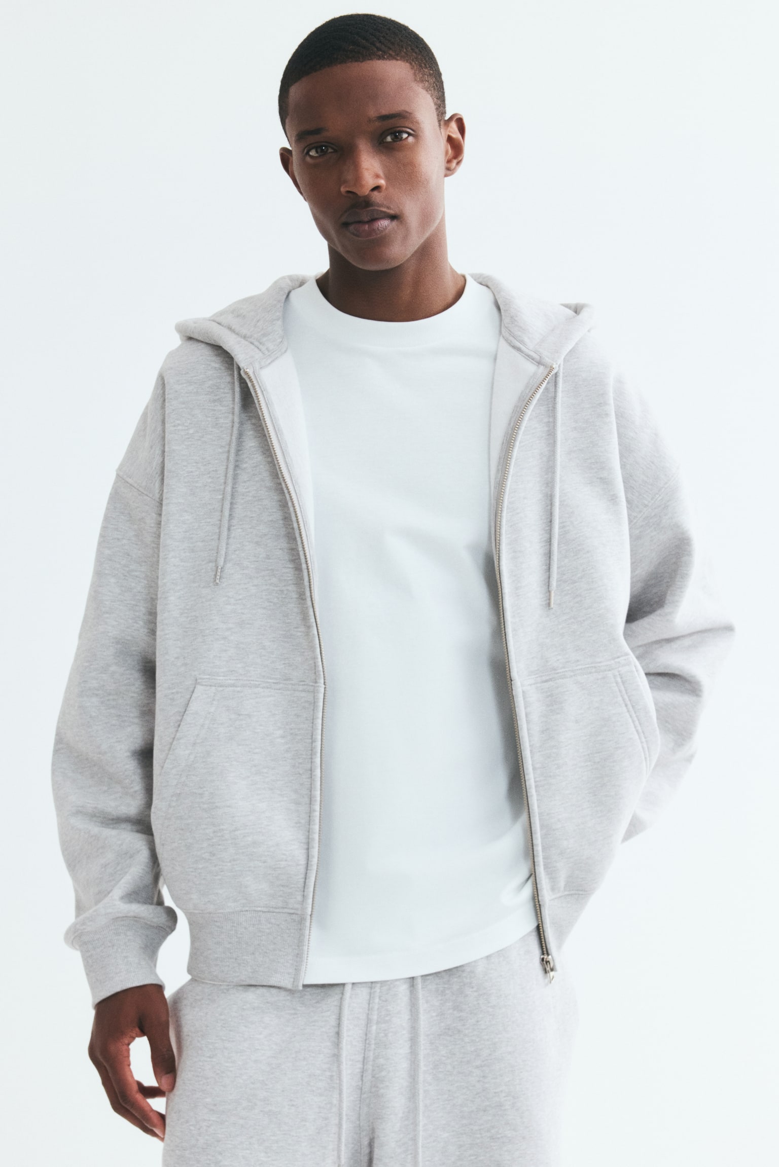 A light grey zip-through hoodie is on display, showcasing its front with a full-zip closure and a jersey-lined drawstring hood. Crafted from sweatshirt fabric, it boasts dropped shoulders, long sleeves with ribbed cuffs, front pockets, and a ribbed hem, offering an oversized, baggy silhouette. - 1