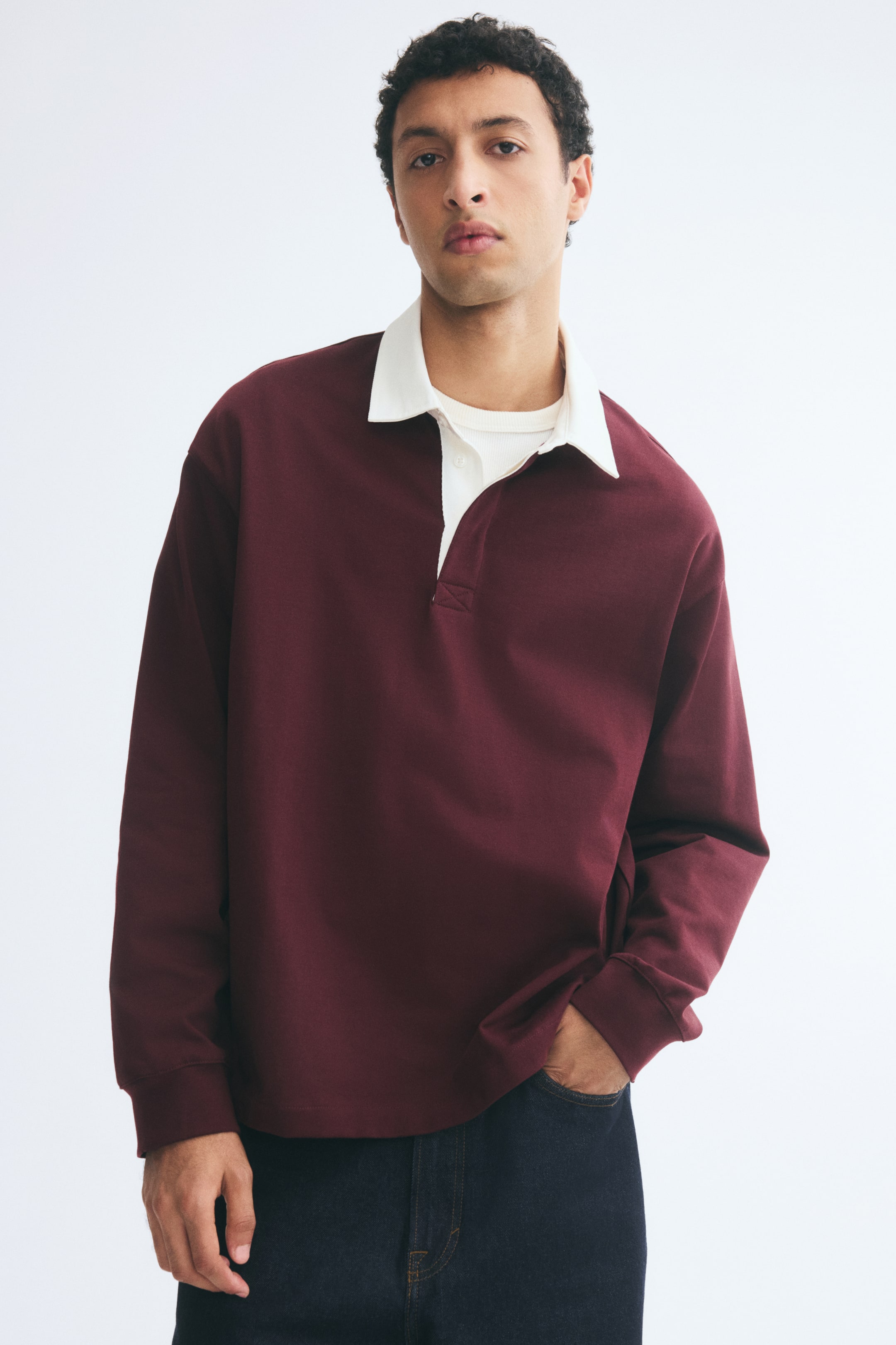 View larger image: A maroon rugby shirt with a contrasting white collar and ribbed cuffs is being worn by the model over a white crew-neck T-shirt. The relaxed-fit shirt is paired with dark jeans, with the model placing one hand in a front pocket.