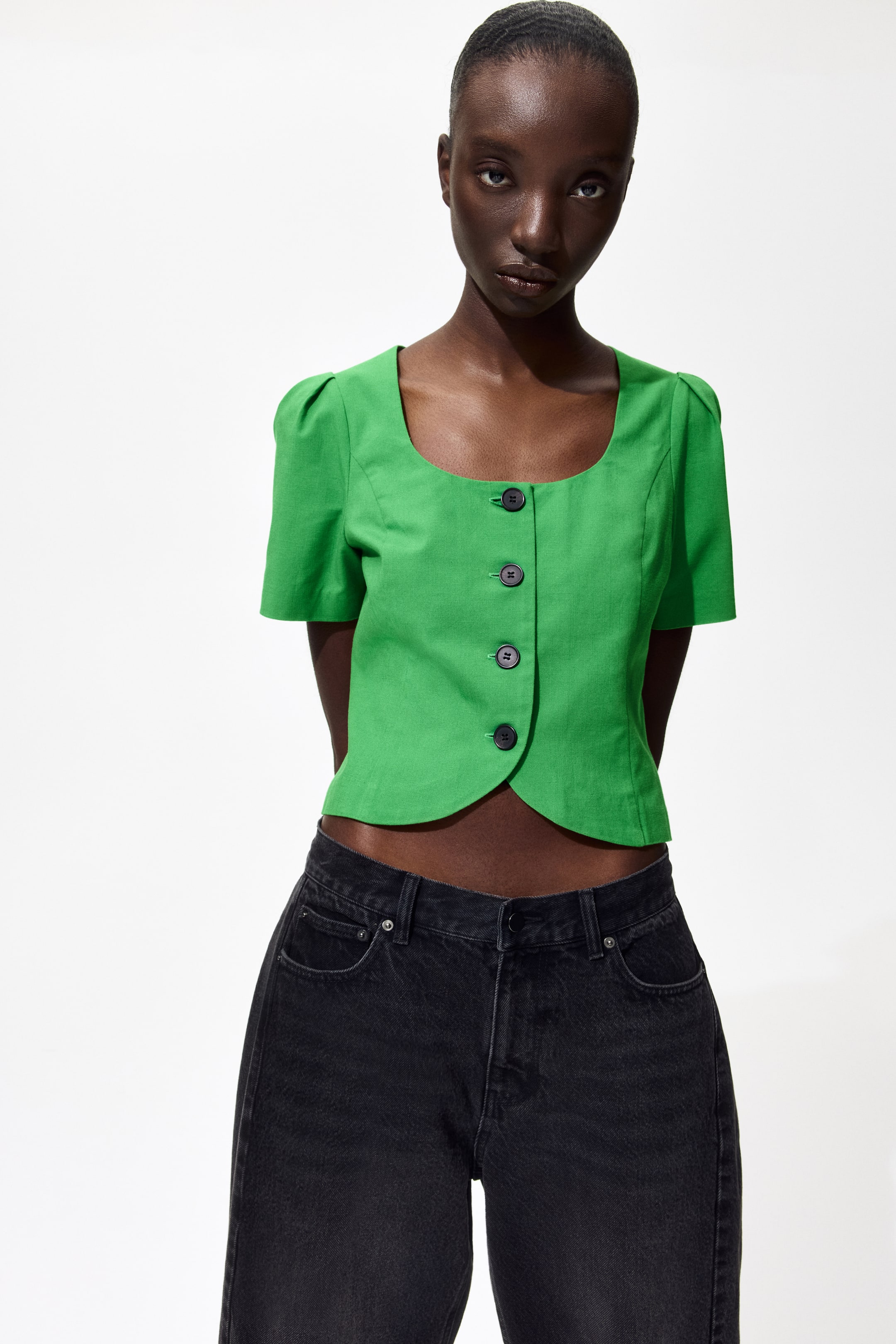 Ver imagem maior: Uma blusa curta, verde e justa com decote redondo, botões escuros na frente e mangas curtas bufantes está a ser usada pela modelo, combinada com jeans escuros.