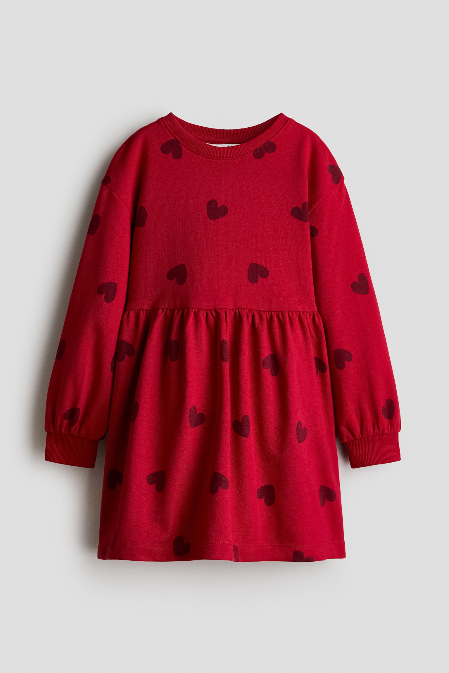 Ein rotes, oversized Fleecekleid mit einem allover dunkelroten Herzmuster ist von vorne zu sehen. Das Kleid zeigt einen runden, gerippten Ausschnitt, Ã¼berschnittene Schultern, lange Ãrmel mit gerippten BÃ¼ndchen und eine geraffte Naht in der Taille.