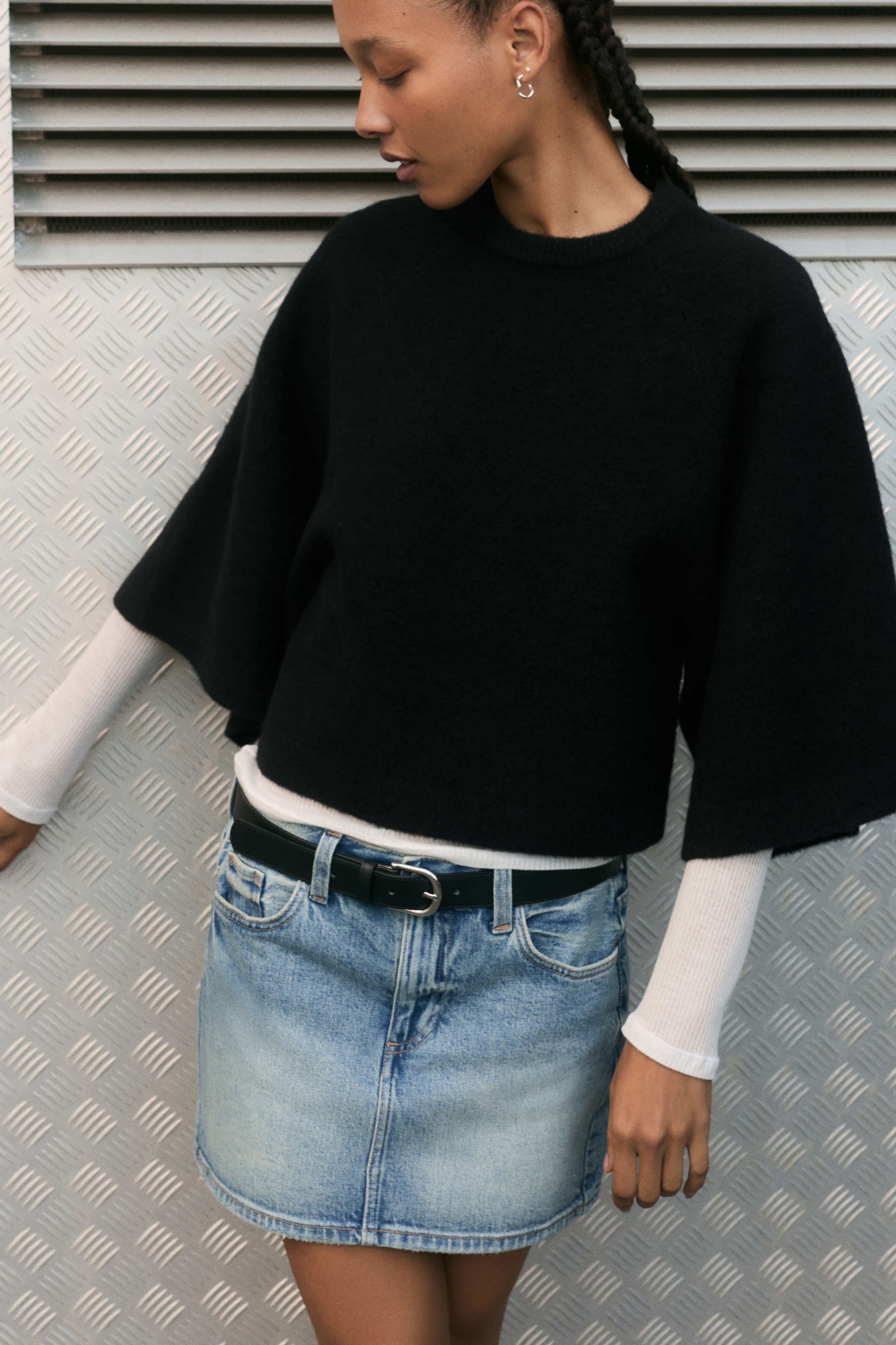 Ver imagen más grande: La modelo lleva un jersey de punto negro de corte relajado, con cuello redondo rematado en canalé y mangas raglán anchas de largo 3/4. El jersey se combina sobre una camiseta blanca de manga larga y con una falda vaquera azul.