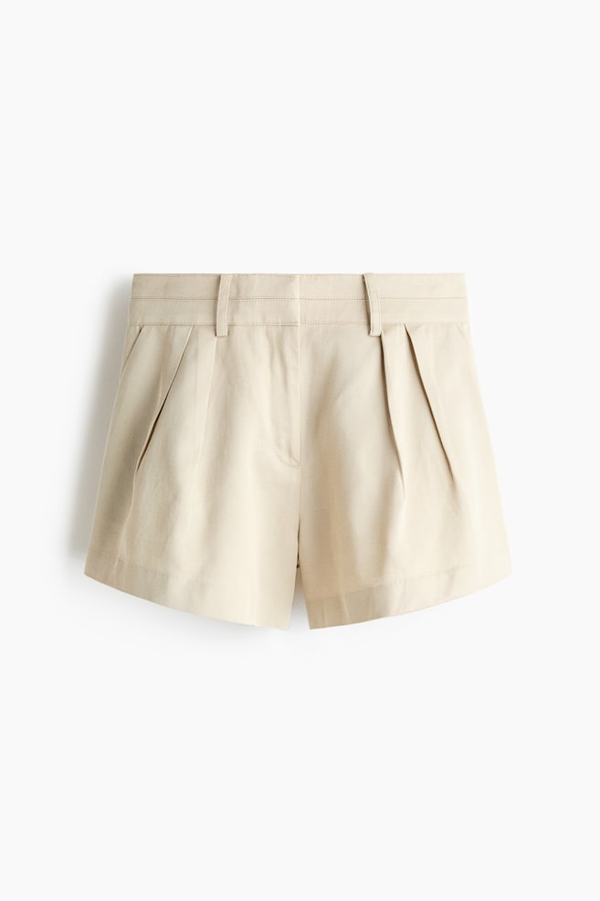 Light beige shorts are presented in a front view, featuring a waistband with belt loops. Two prominent forward-facing pleats detail each front side, with side slash pockets also visible. - 1
