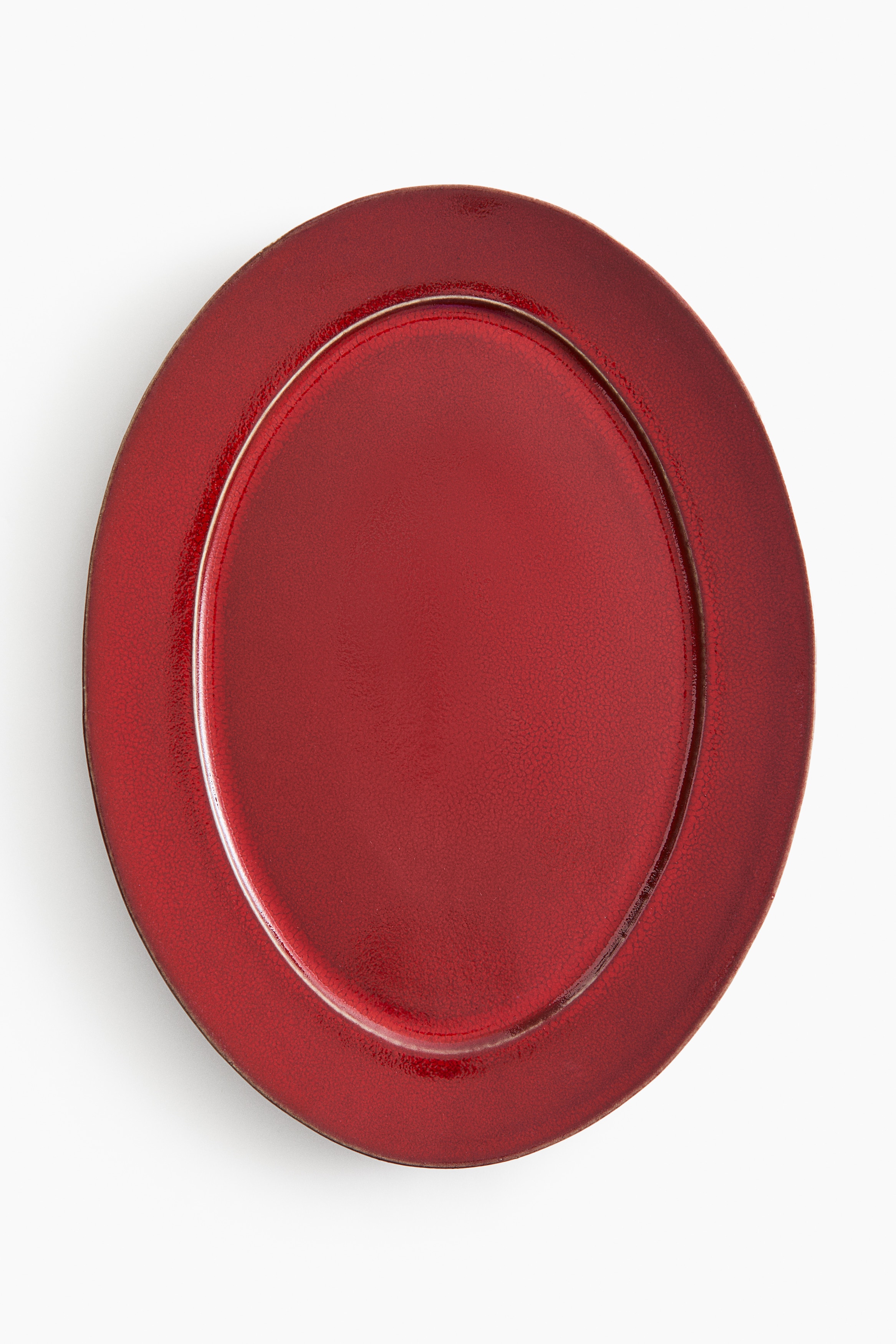 Un plat ovale en céramique, d’un rouge profond et brillant, est présenté en vue de dessus, avec un fin liseré clair soulignant le bord de sa base intérieure. Une petite grappe de raisin rouge repose au centre du plat, mettant en valeur sa finition lisse et réfléchissante.