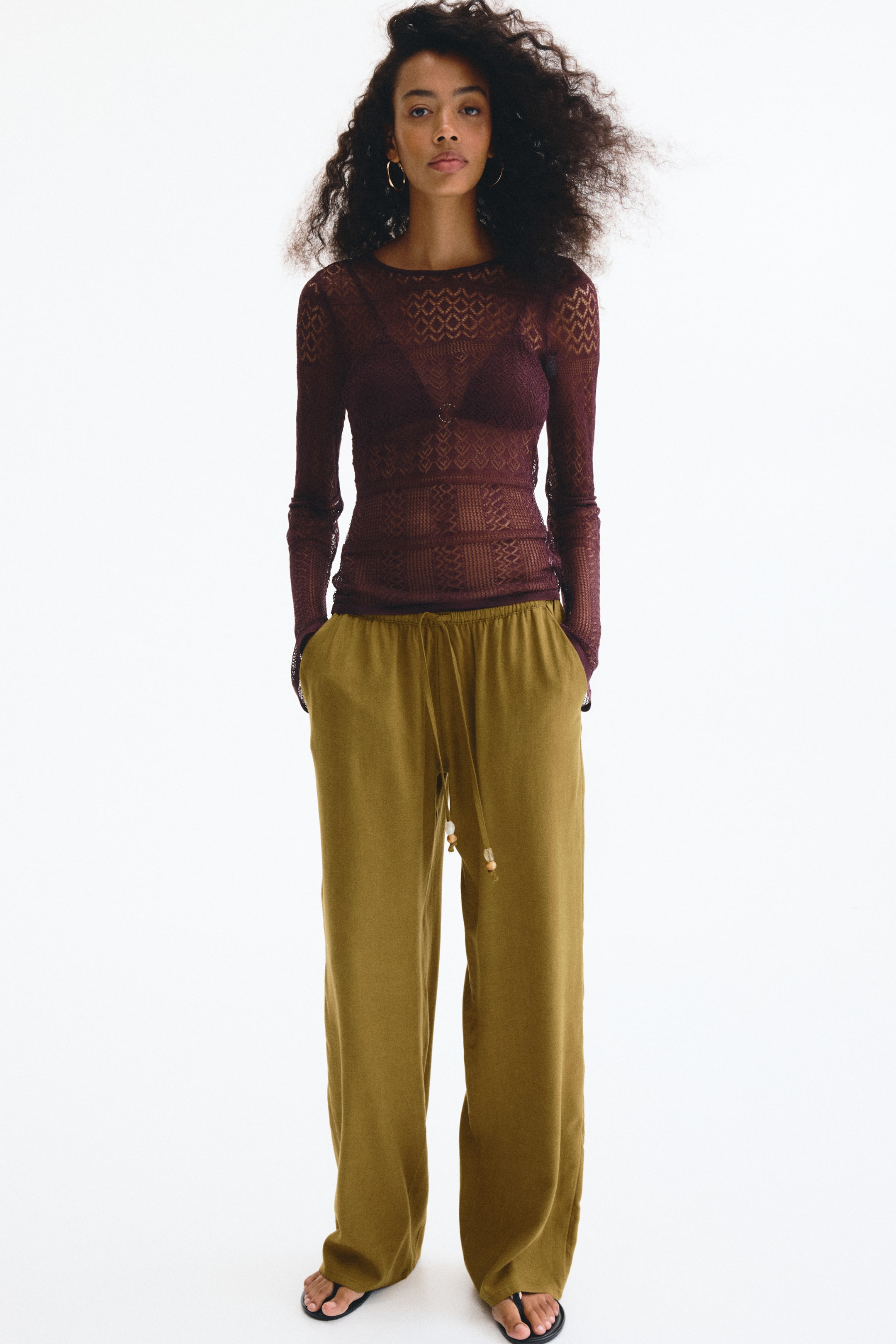 View larger image: A sheer, long-sleeved plum top with a geometric diamond knit pattern is being worn over a dark bra. It is being paired with olive green wide-leg pants featuring an elastic drawstring waistband with beaded detailing and side pockets.