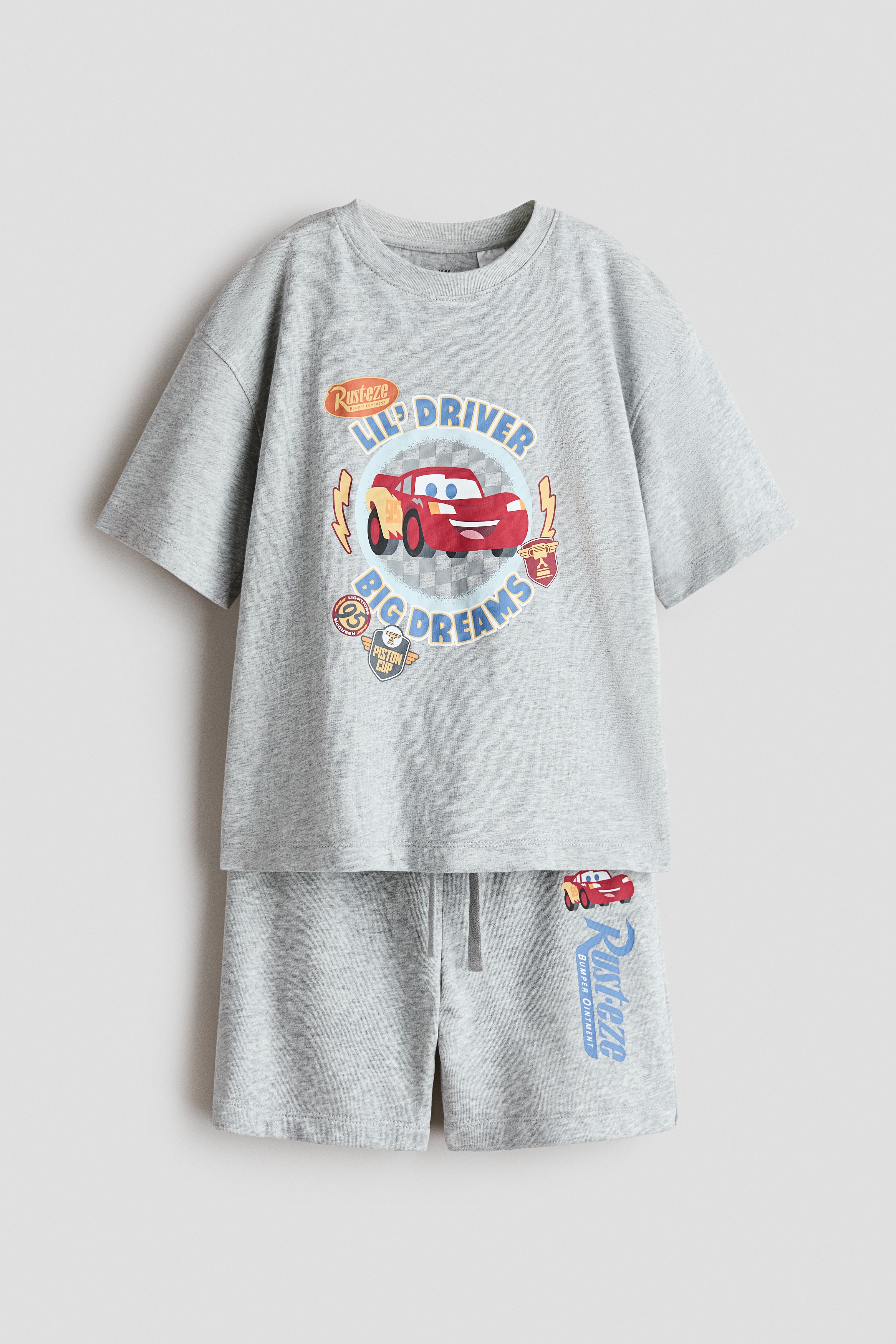 A light grey jersey T-shirt featuring a colourful graphic print of a red race car and the text "LIL' DRIVER BIG DREAMS" is being worn by the model. The model is posing with their hands placed behind their head, highlighting the T-shirt's round neckline and relaxed fit.
