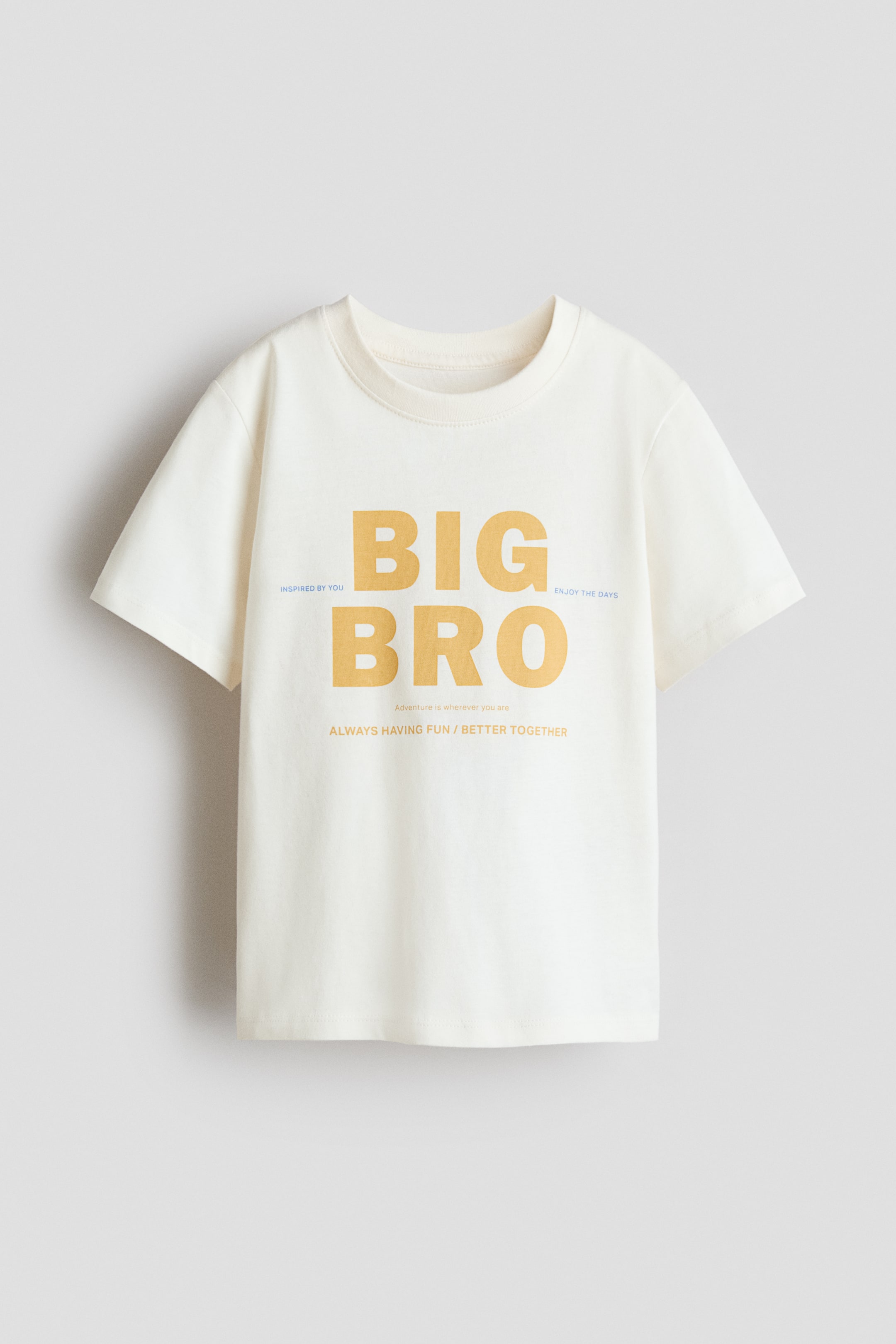 View larger image: A cream T-shirt is shown from the front, featuring a ribbed round neckline, dropped shoulders, short sleeves and a straight-cut hem. The front bears "BIG BRO" in large gold lettering, accompanied by smaller blue and gold text.