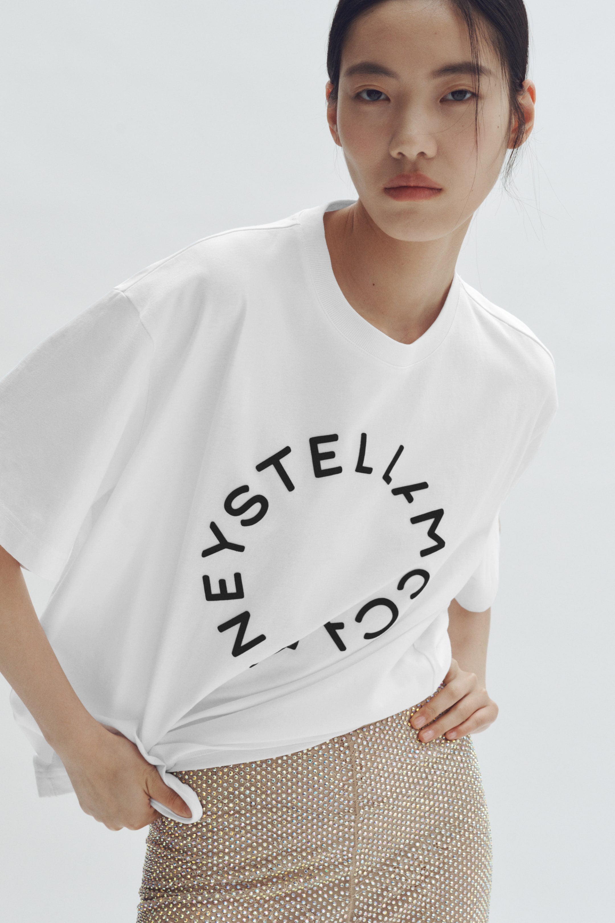 Visa större bild: En vit t-shirt visar en stor cirkulär svart grafik med text arrangerad i en cirkel på framsidan, med ribbad rund halsringning och korta ärmar. Ena sidan av t-shirtens fåll hålls upp, vilket visar dess avslappnade passform.