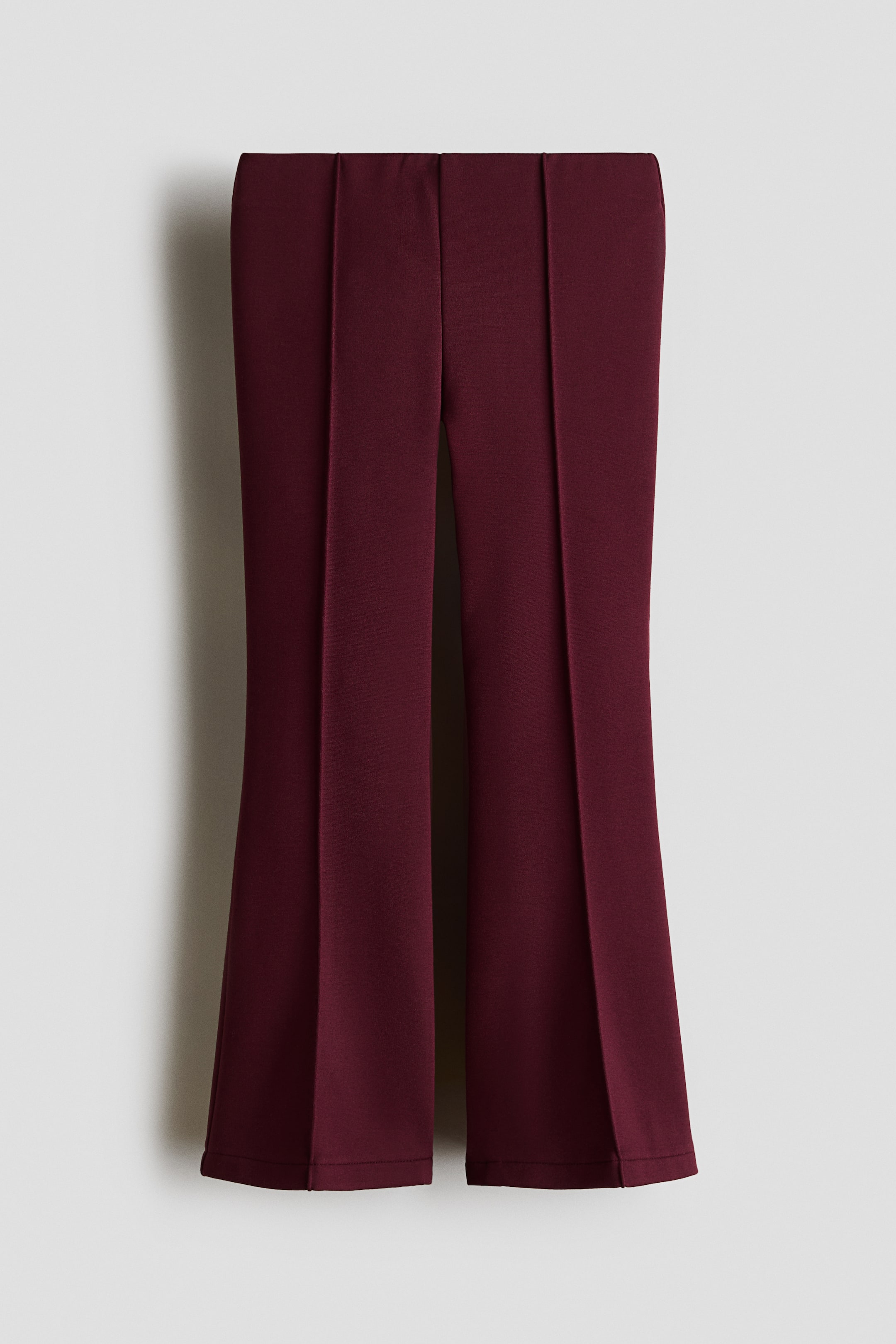 View larger image: These flared burgundy leggings in heavyweight ponte fabric feature a flat, concealed waistband and sewn-in creases running down the centre of each leg. The legs flare outward towards the hem to create a structured silhouette in a front view.