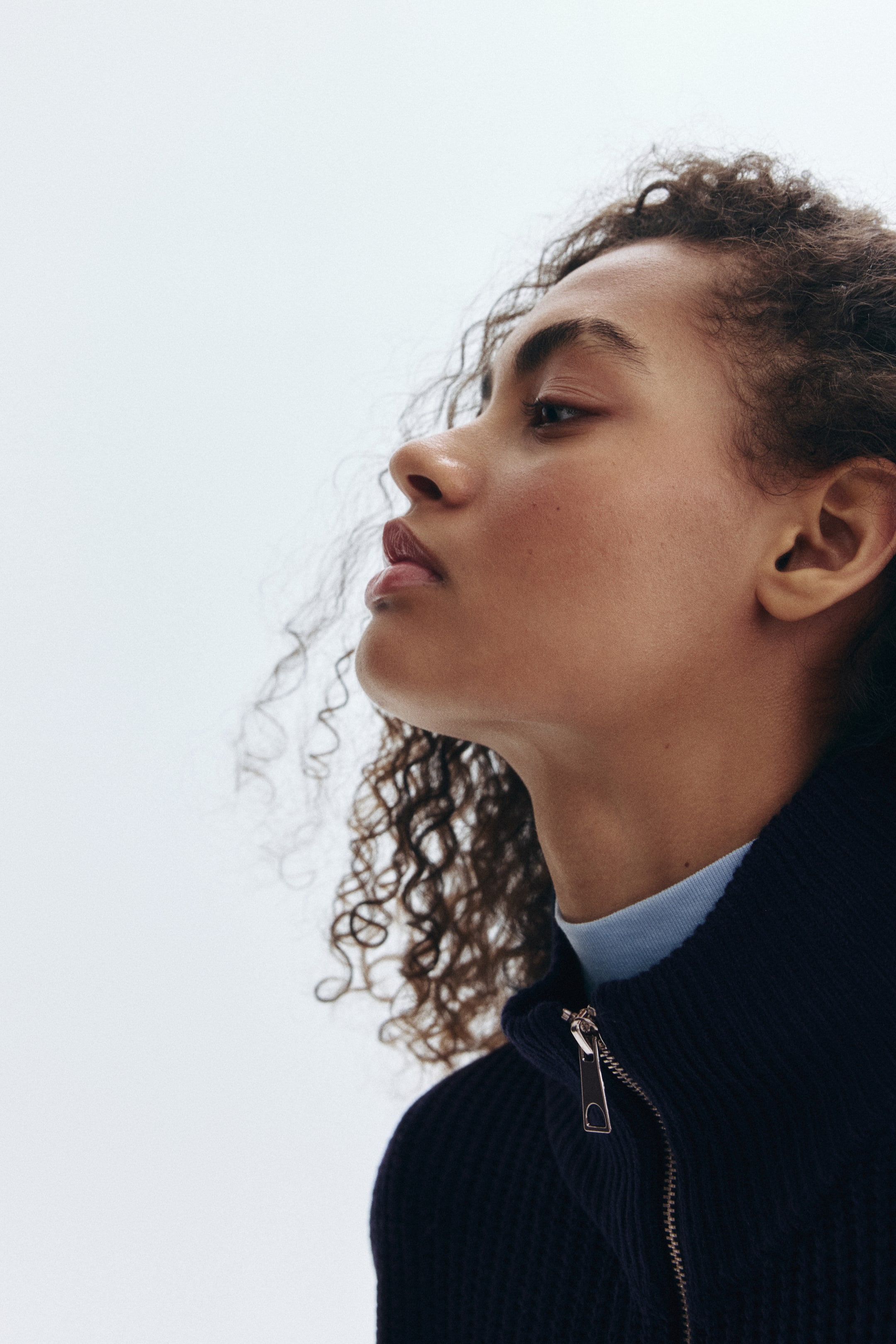 View larger image: A dark navy, ribbed, full-zip sweater is being worn by the model. The sweater is featuring a high collar and a silver-toned zipper pull, with a light blue top visible underneath.