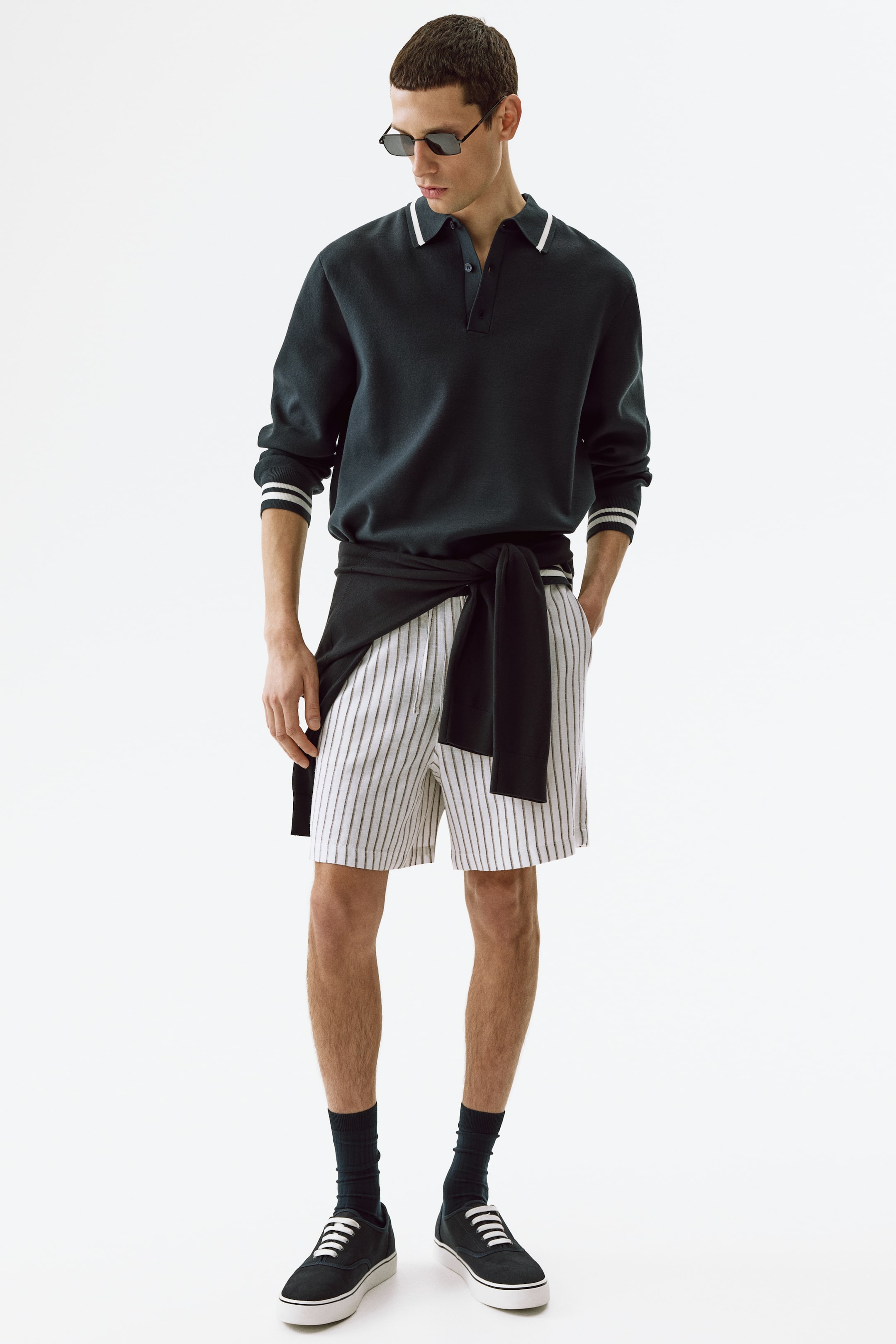 Ingrandisci l'immagine: Pantaloncini ariosi in lino e viscosa con righe verticali bianche e scure indossati dalla modella. I pantaloncini presentano un laccio in vita, una patta finta e tasche laterali diagonali, una delle quali ospita la mano sinistra della modella. Un maglione scuro è anche annodato intorno alla vita della modella sopra i pantaloncini.