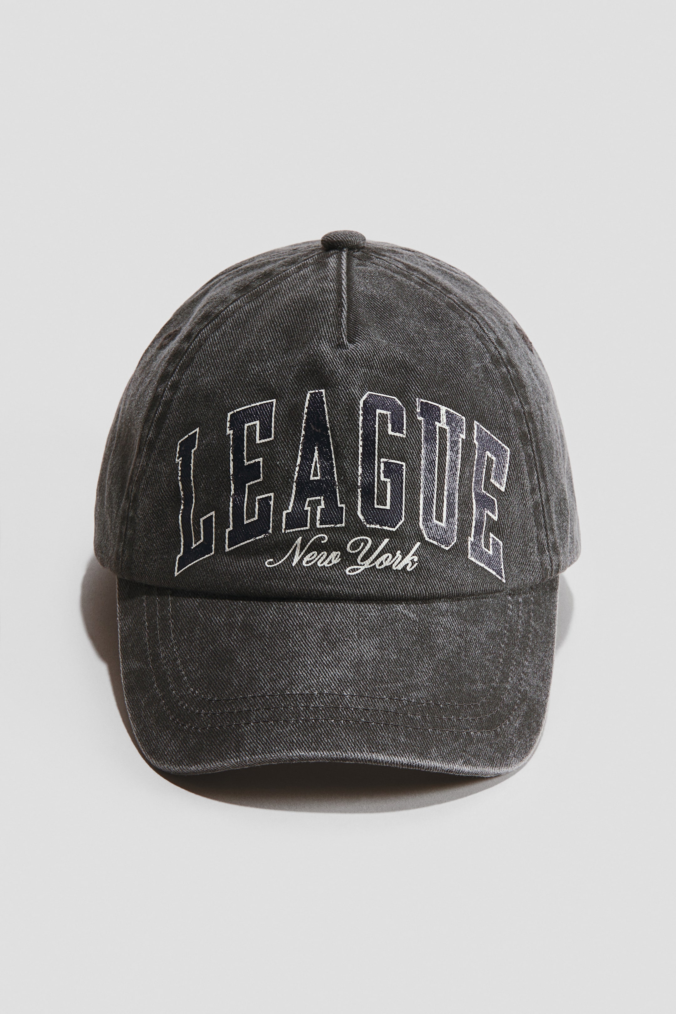 View larger image: The image displays a dark grey cotton twill cap positioned front-facing, showcasing an arched "LEAGUE" design in dark blue accented by a white outline, with "New York" elegantly scripted in smaller white cursive beneath. The cap is equipped with embroidered eyelets, an adjustable strap featuring a hook and loop fastener at the back, and a cotton sweatband.